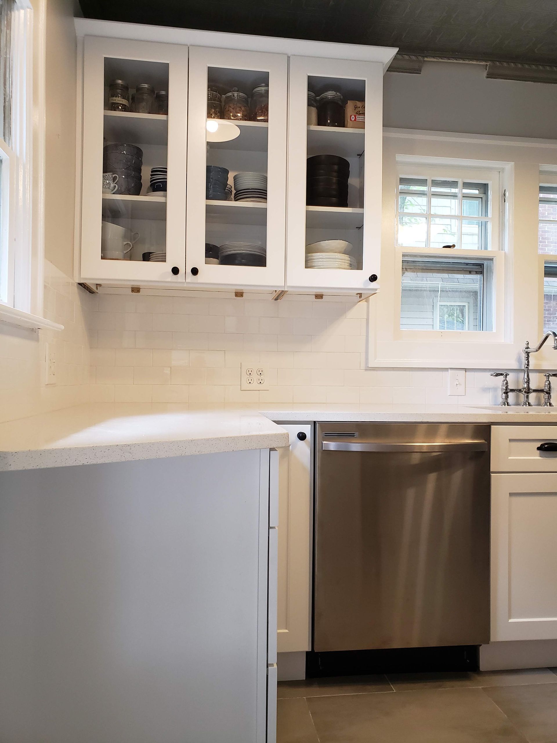 Bright white kitchen with glass-front cabinets, dishwasher, sink, and window above the counter.