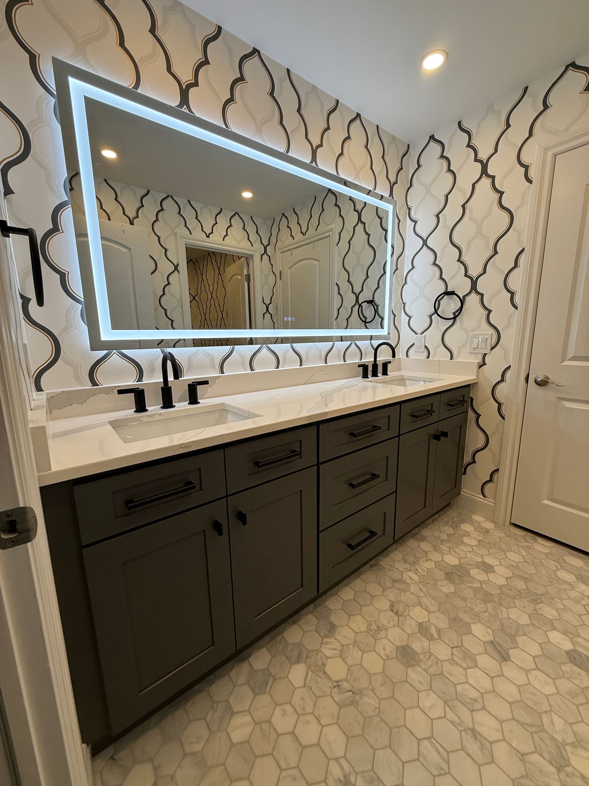 Modern bathroom vanity with backlit mirror, dark cabinets, white countertop, and patterned wallpaper