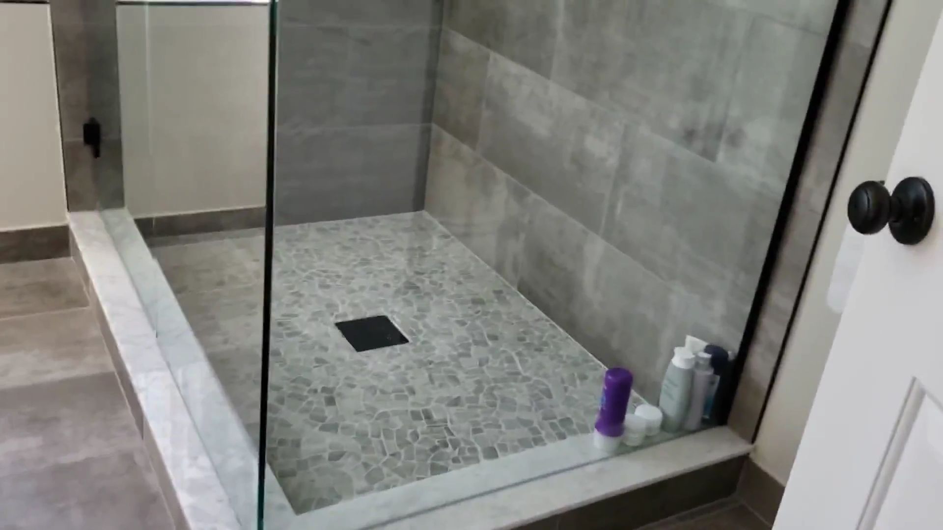 Modern walk-in shower with glass doors, gray tile, and a black floor drain.