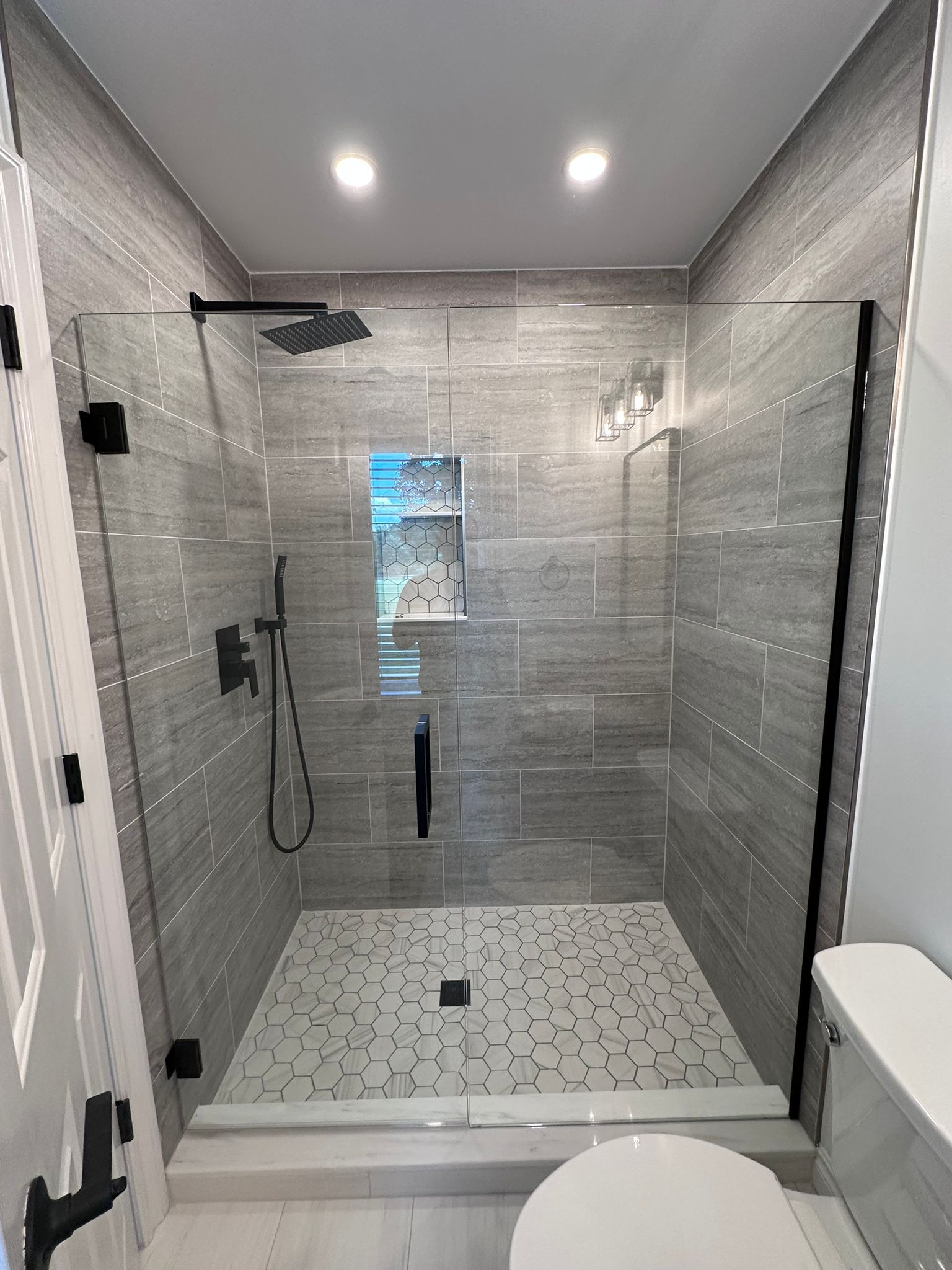 Modern gray-tiled walk-in shower with glass door, black fixtures, and white hexagon floor tiles