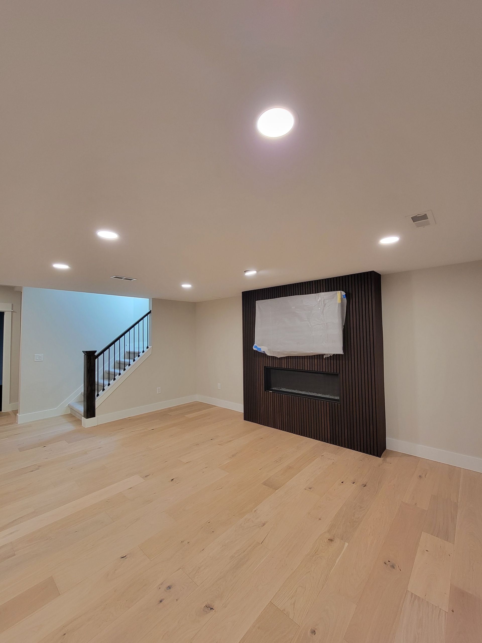 Empty living room with light wood floors, recessed lights, staircase, and a wall-mounted TV under a striped accent wall.