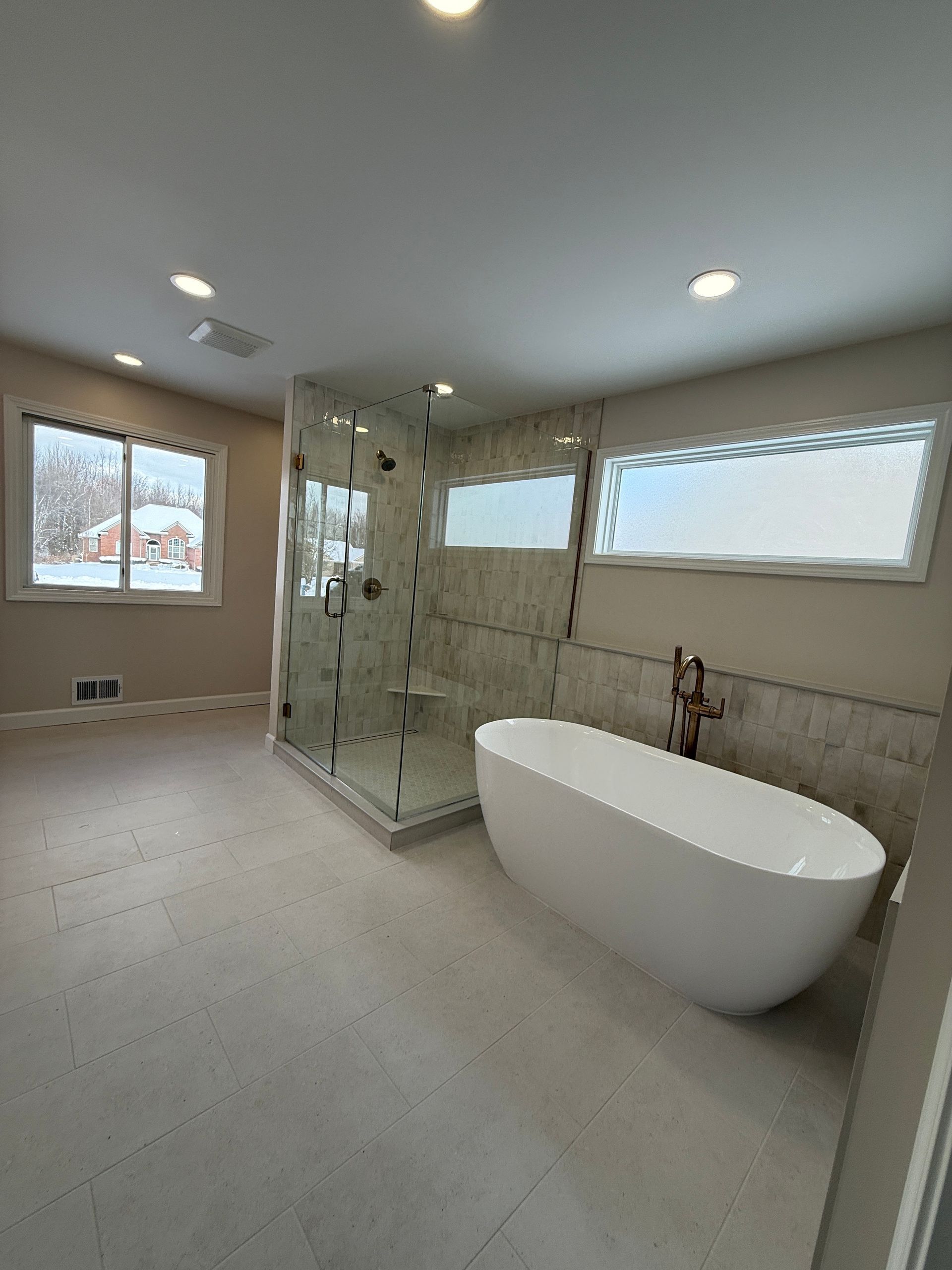Modern bathroom with freestanding tub, glass shower, windows, and light tile flooring