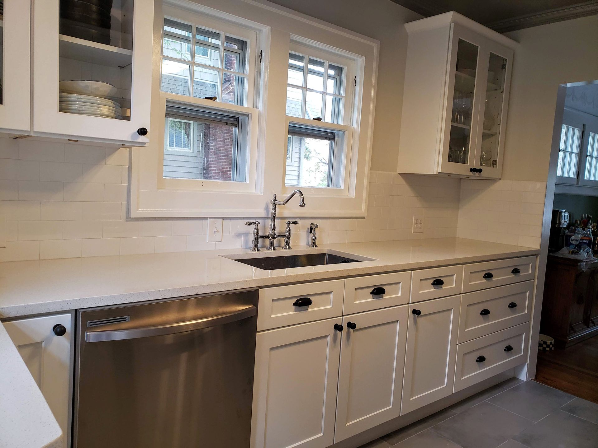 Bright white kitchen with marble countertops, stainless dishwasher, sink under windows, and built-in cabinets.