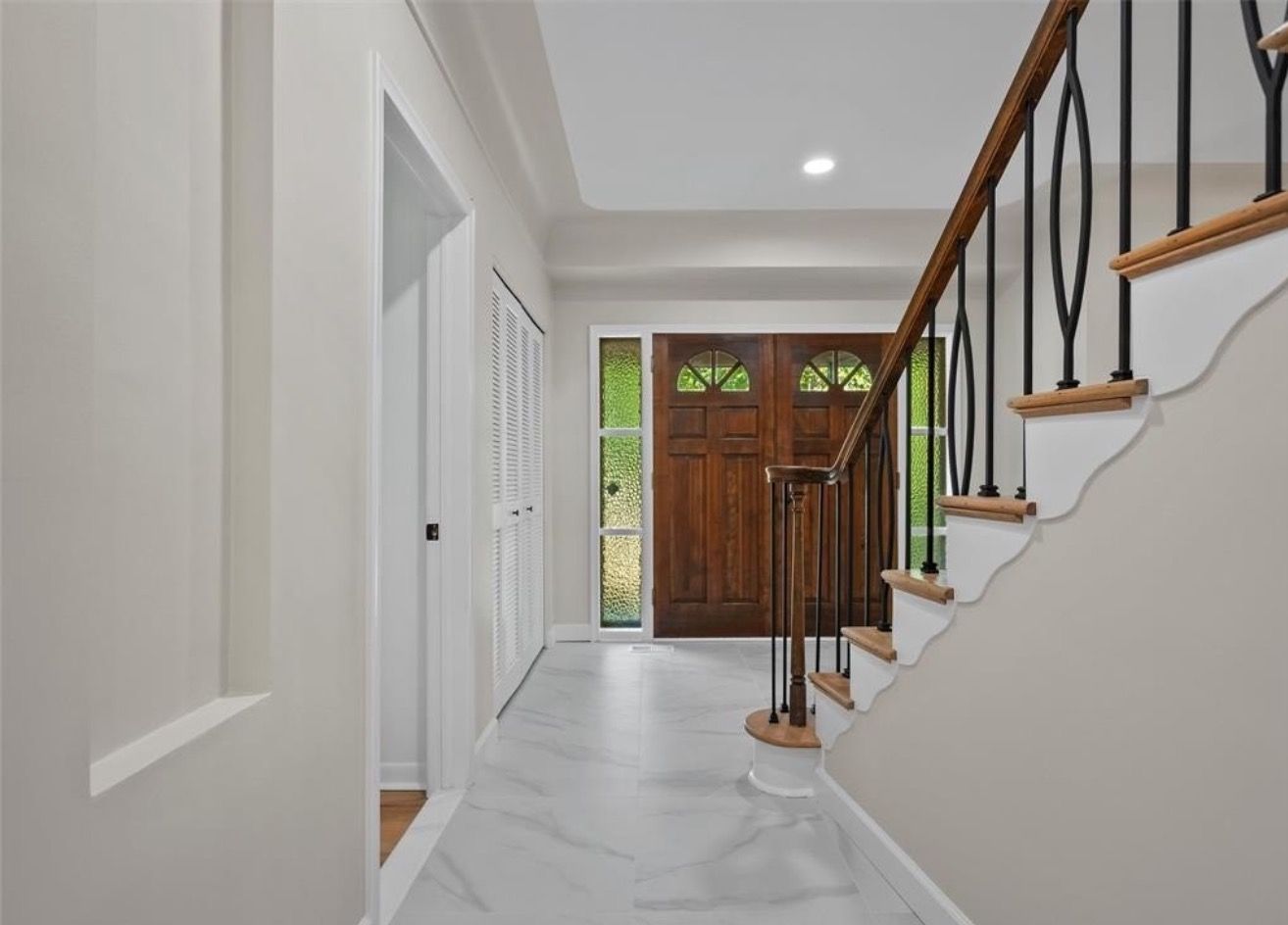 Bright hallway with white walls, marble floor, stairs, and a wooden front door at the end.