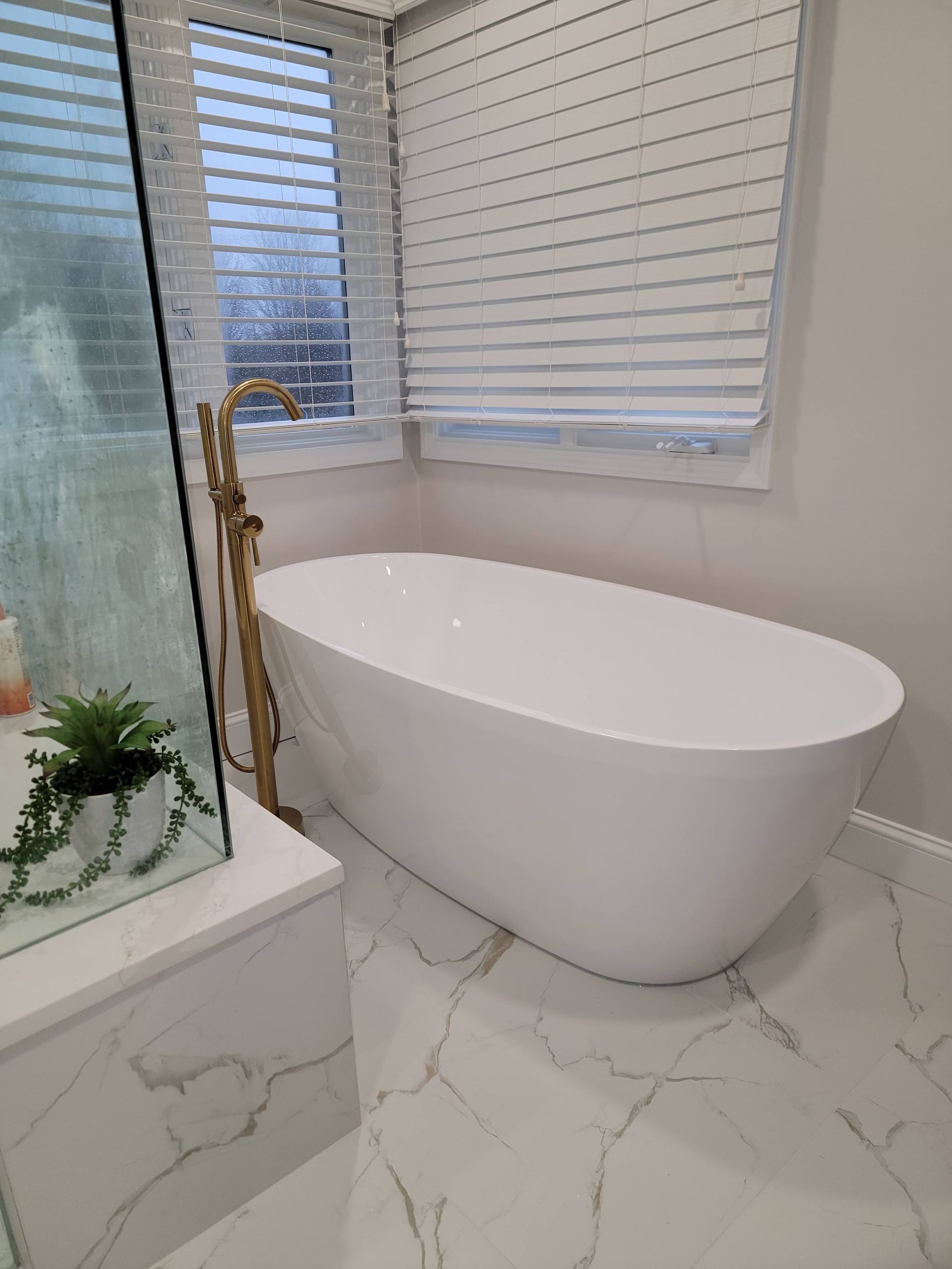 White freestanding bathtub beside a window with blinds in a marble-tiled bathroom