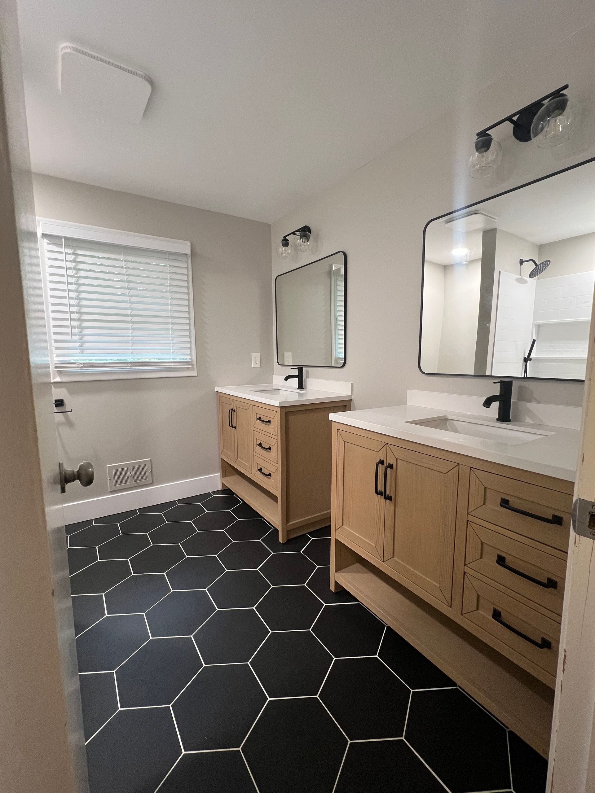 Bright bathroom with black hex tile floor, double vanity, mirrors, and a window with blinds.