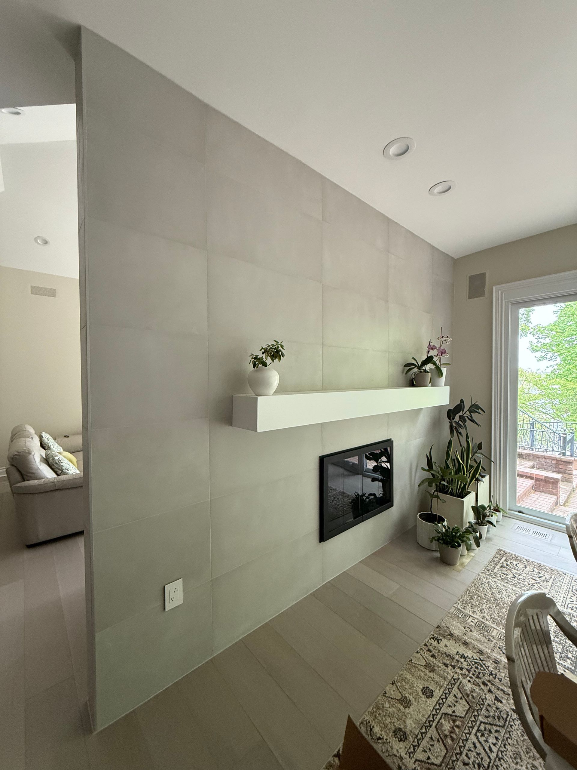 Modern living room with a tiled fireplace wall, floating shelf, and large window overlooking a patio.