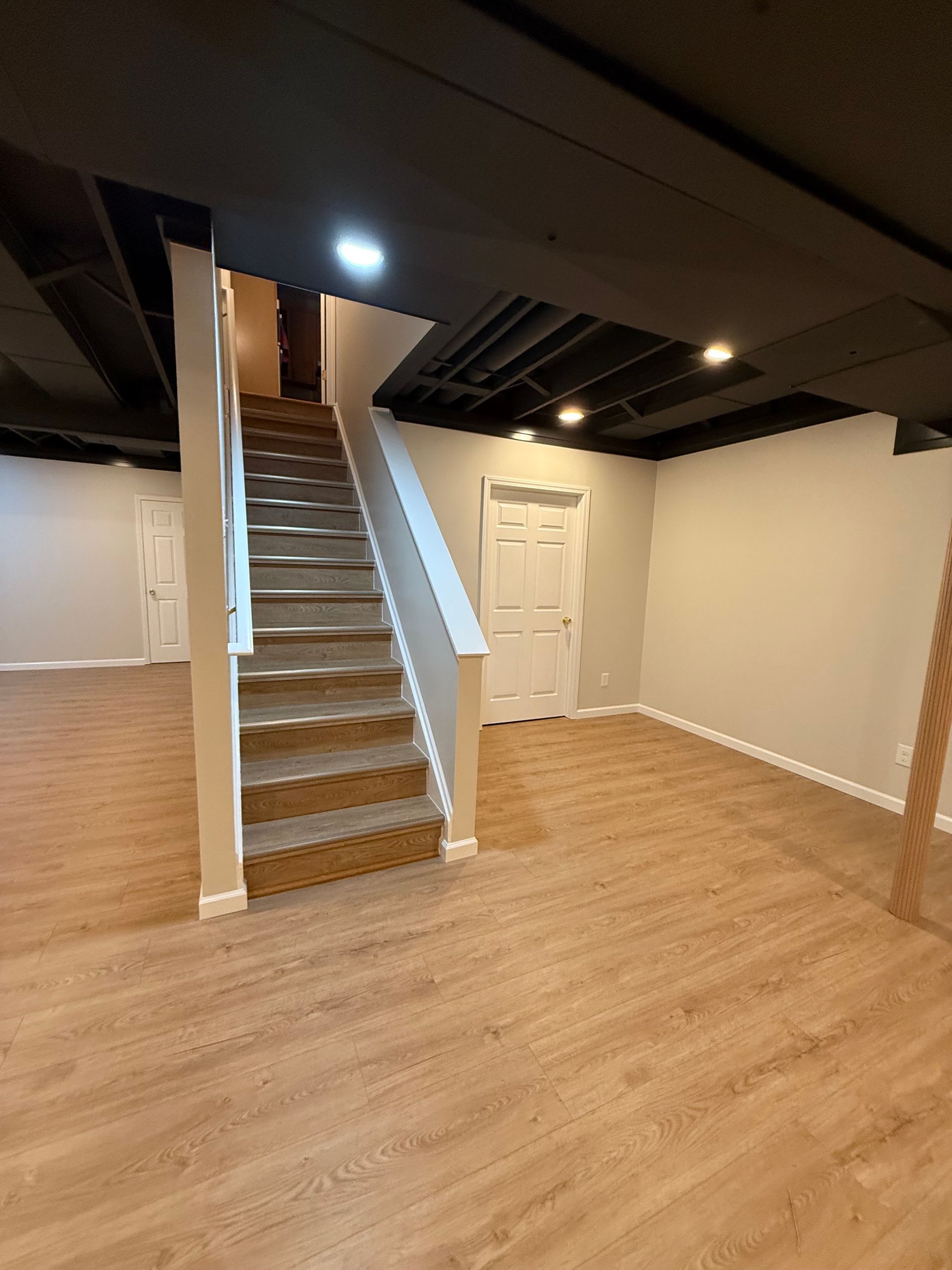 Empty basement room with wood floor, central staircase, white walls, and a door under exposed ceiling beams