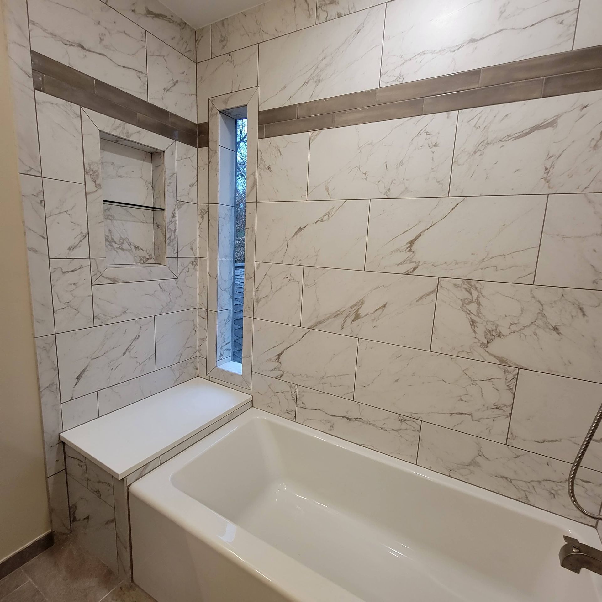 Small modern bathroom with a white bathtub, marble-look tile walls, and a narrow window beside the tub