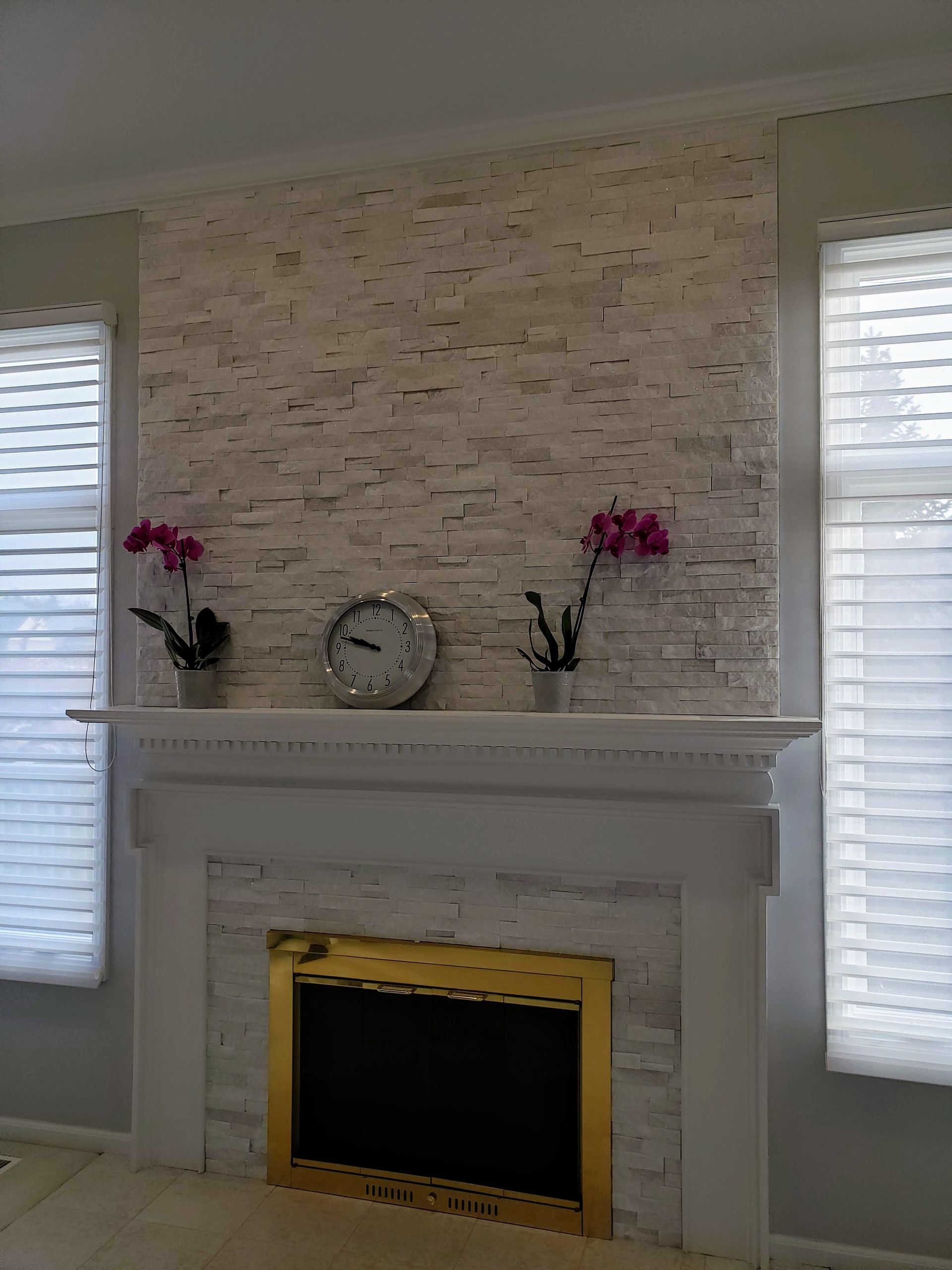 White fireplace with stone surround, two pink flowers, a clock, and window shutters on both sides.