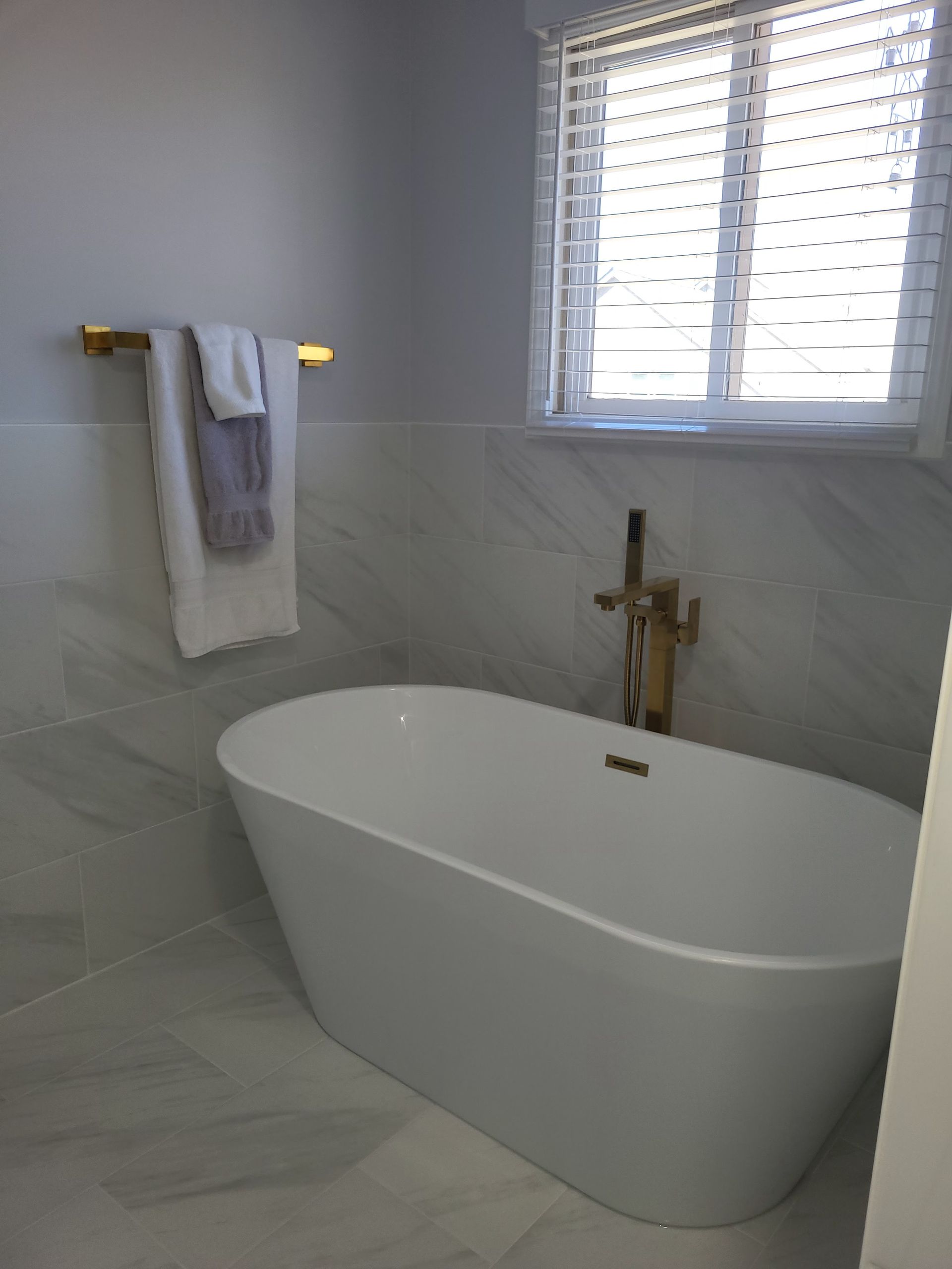Modern bathroom with freestanding white tub, gray marble walls, and window blinds