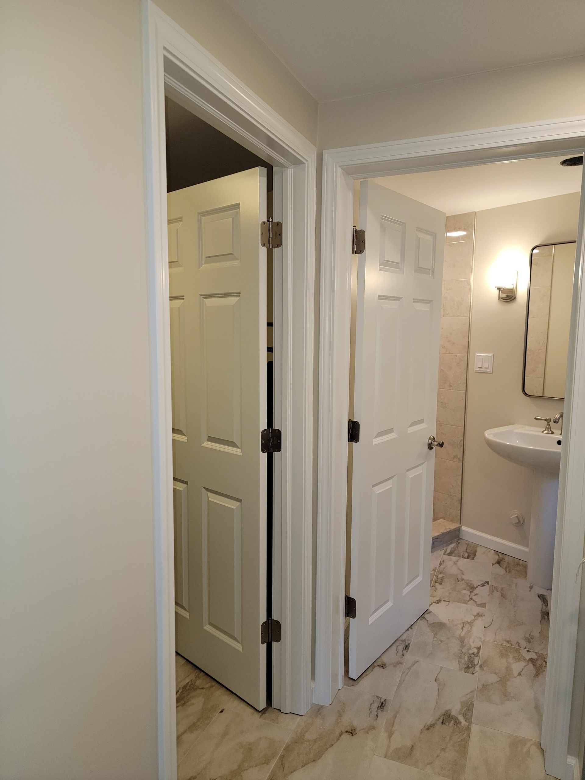 Narrow hallway with open white doors leading to a bright bathroom with a sink and marble-patterned floor.