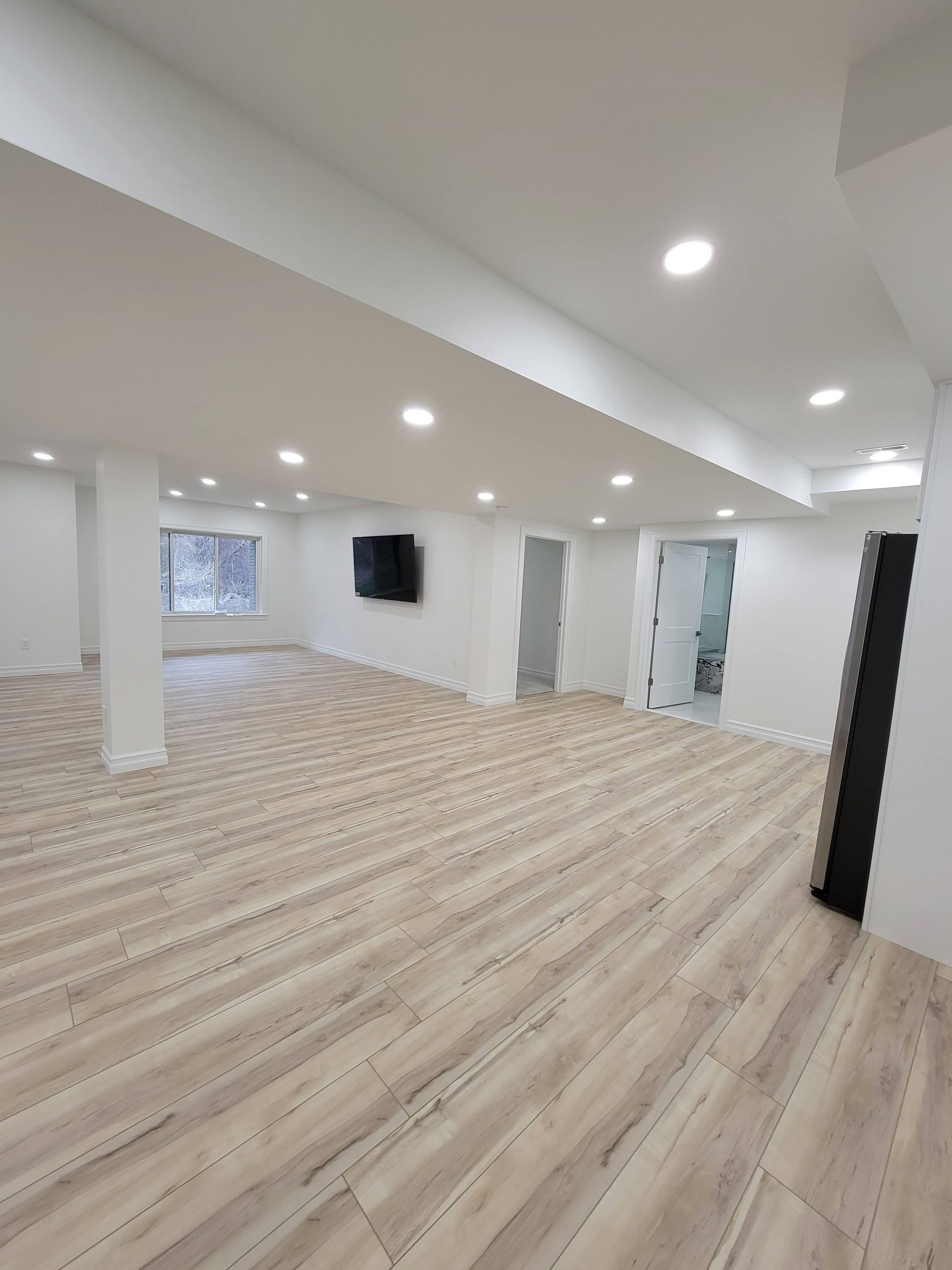 Empty modern room with light wood floors, recessed lights, white walls, and a small wall-mounted TV.