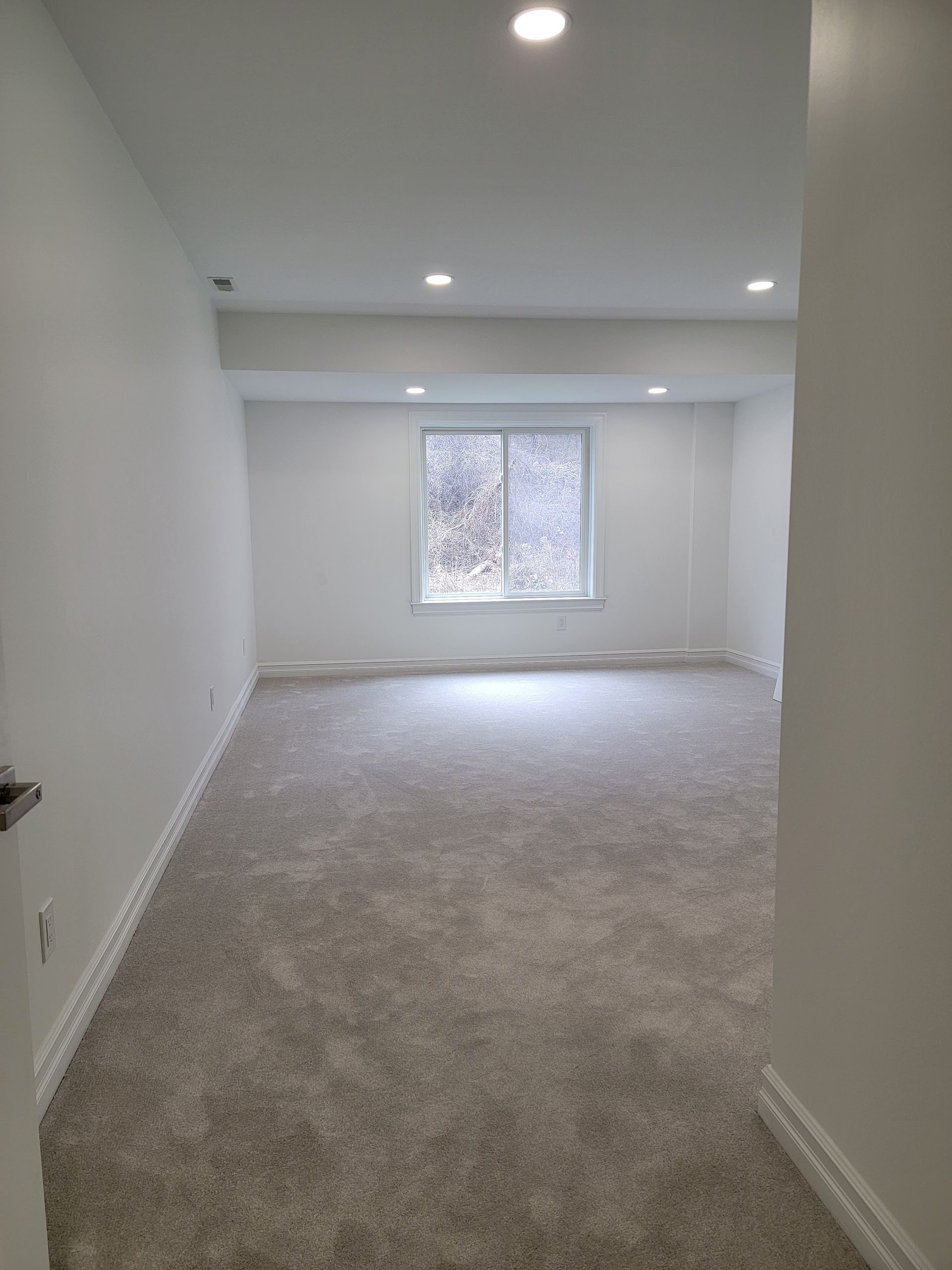 Empty carpeted room with white walls, recessed lights, and a window at the far end.