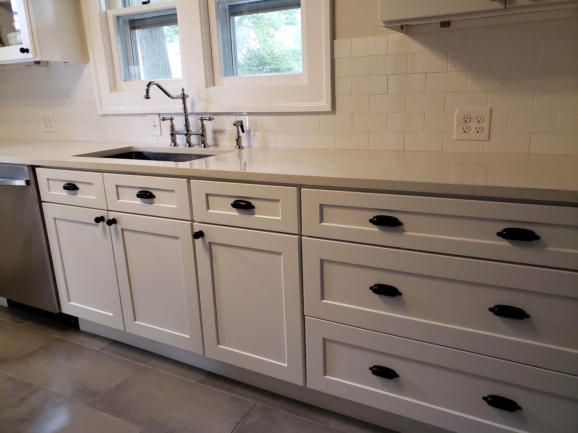 White kitchen with sink, window, and light cabinets with black handles and wood-look floor.