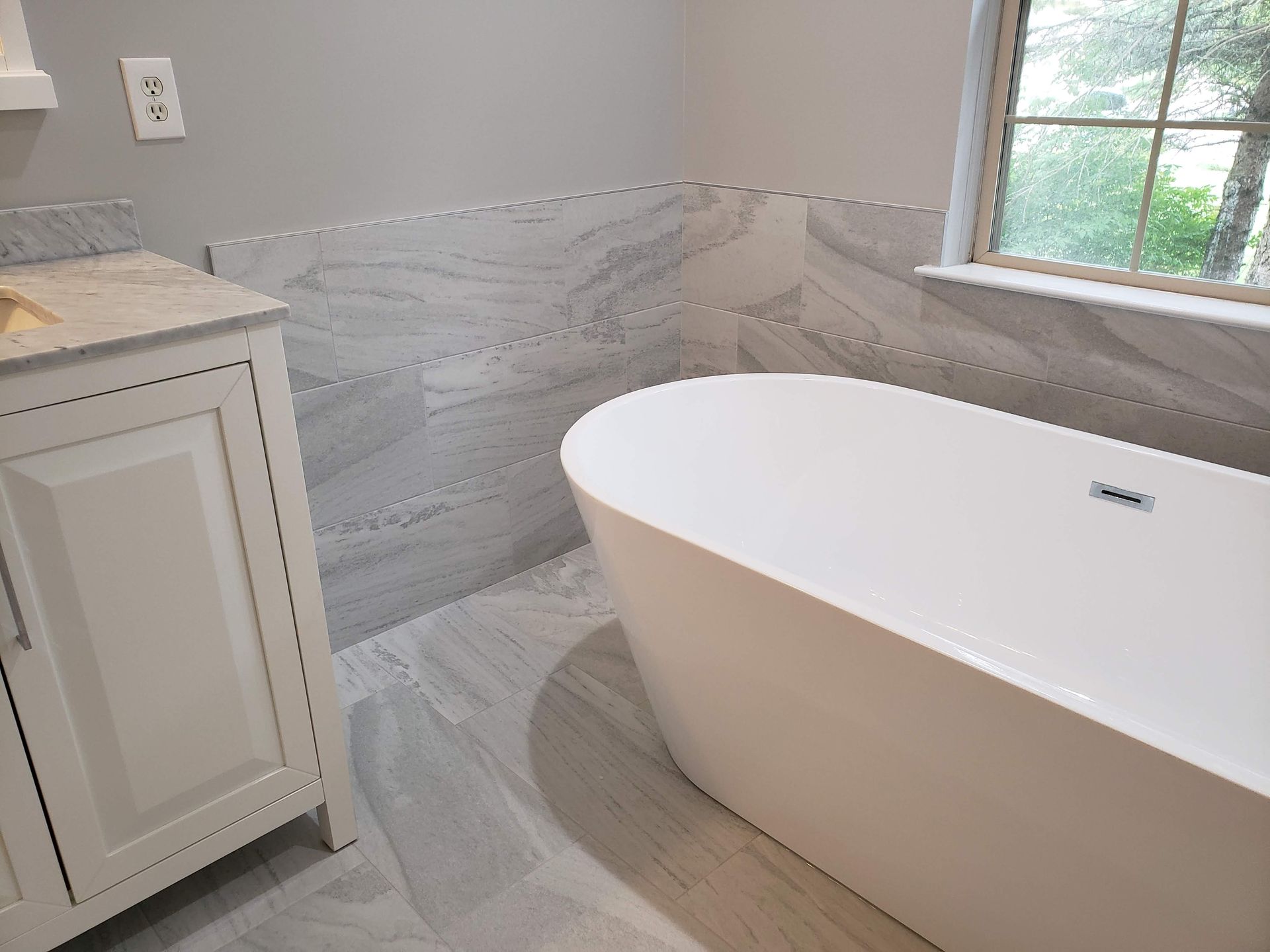 Modern bathroom with a white freestanding bathtub, gray marble tile, and a vanity by a window