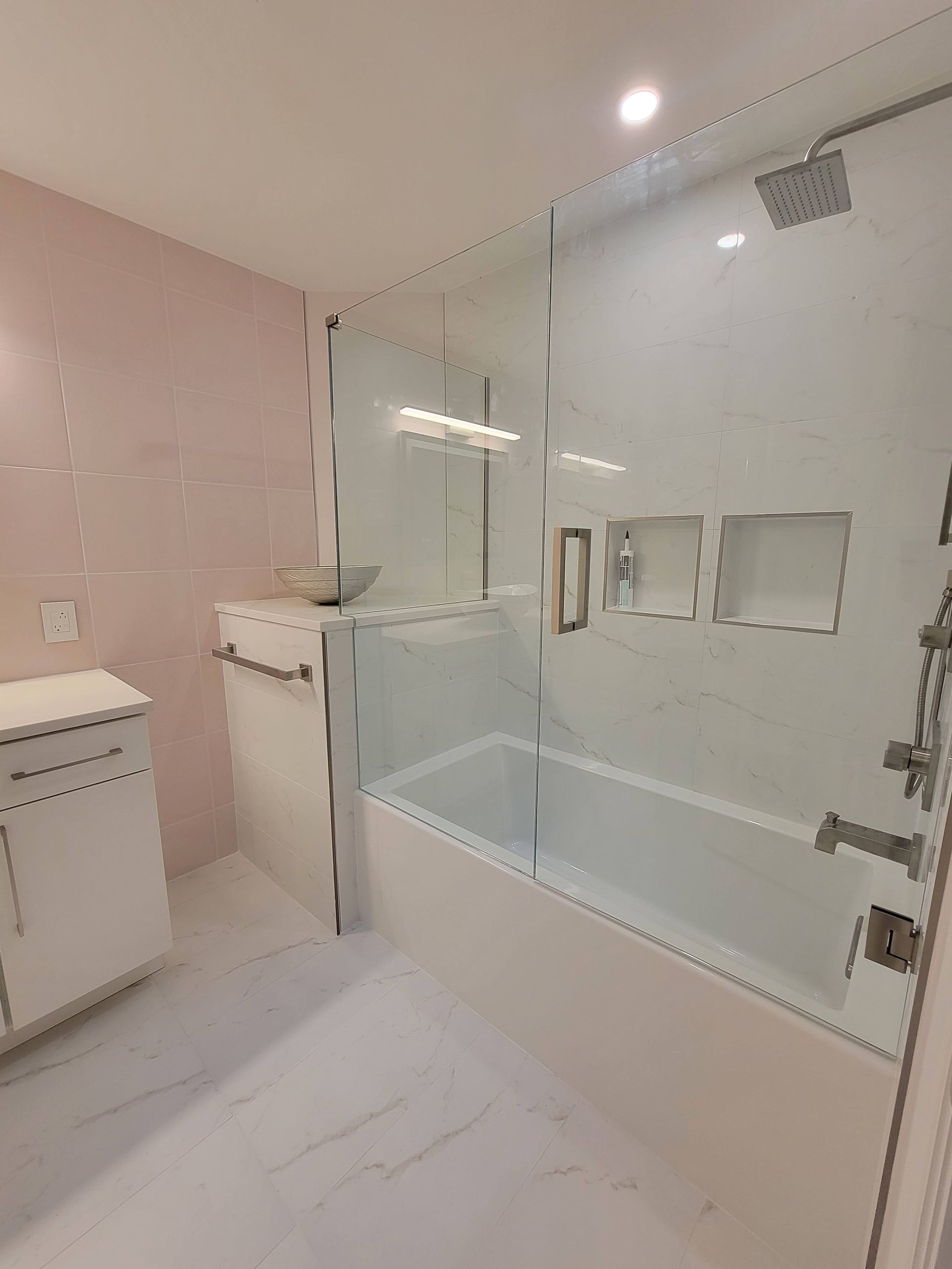 Modern bathroom with white tub shower, glass partition, pink walls, and light tile floor