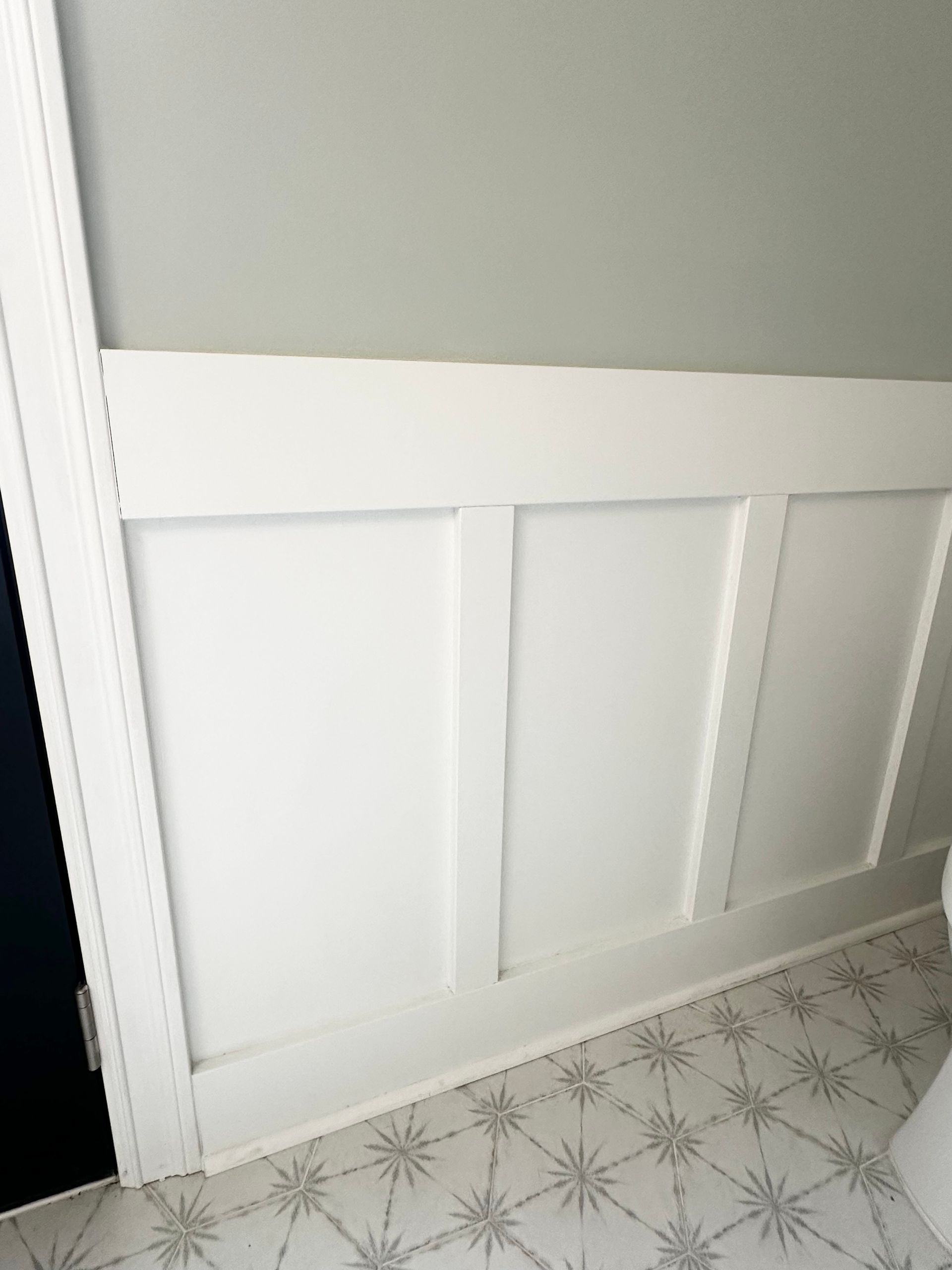 White paneled wainscoting along a light gray wall above patterned tile flooring