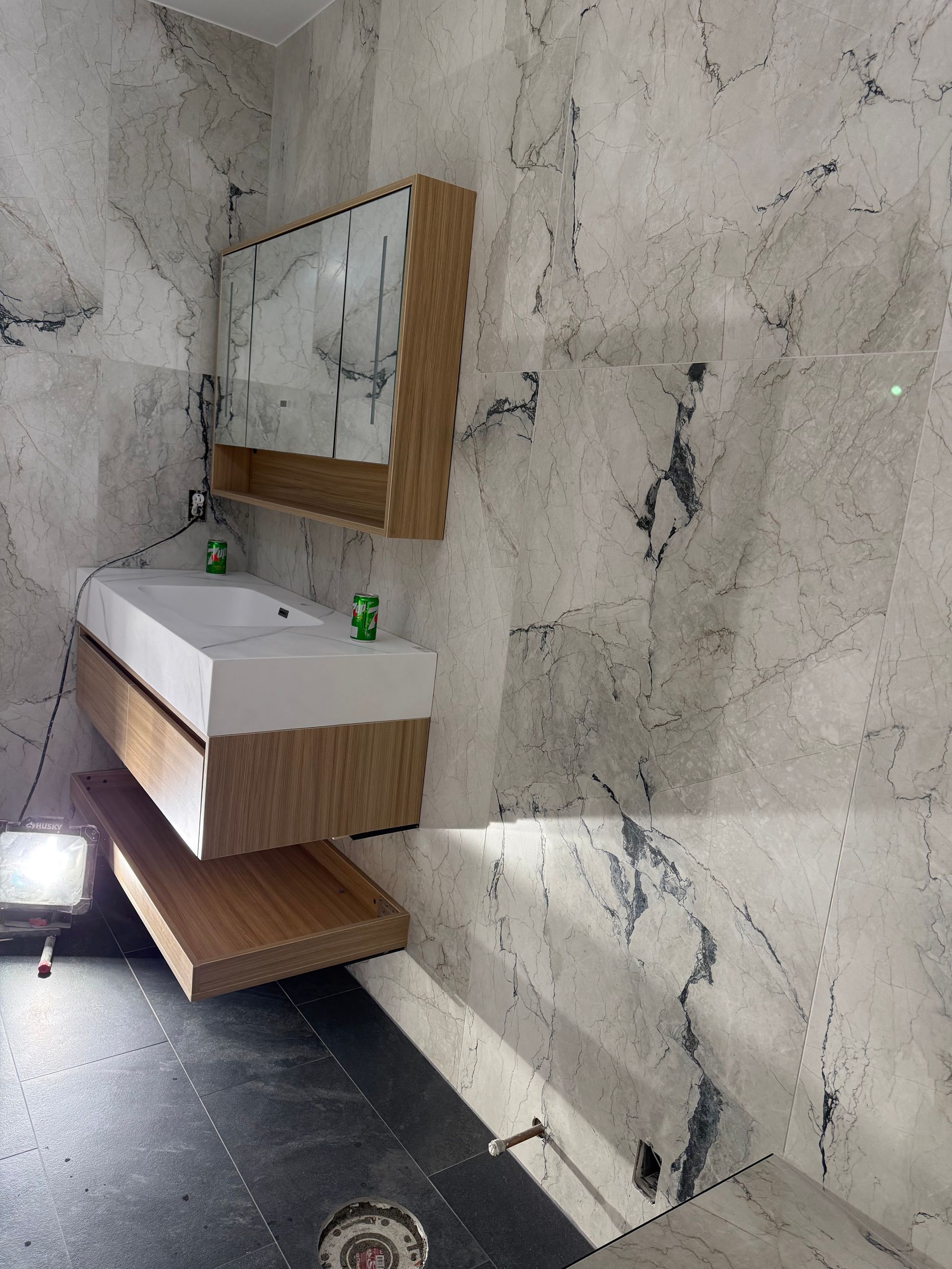 Modern bathroom vanity with wood sink cabinet, mirror, marble-pattern walls, and dark floor lighting.