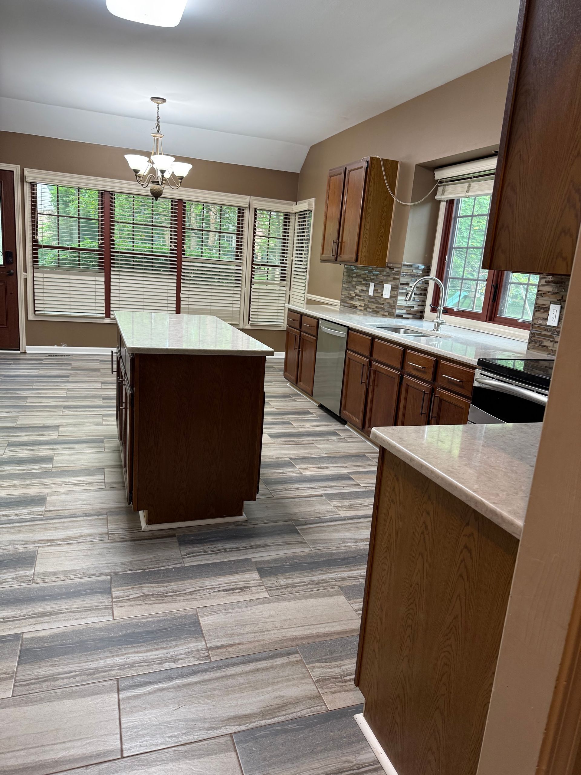 Empty kitchen with wood cabinets, granite countertops, island, and gray plank flooring, lit by windows.