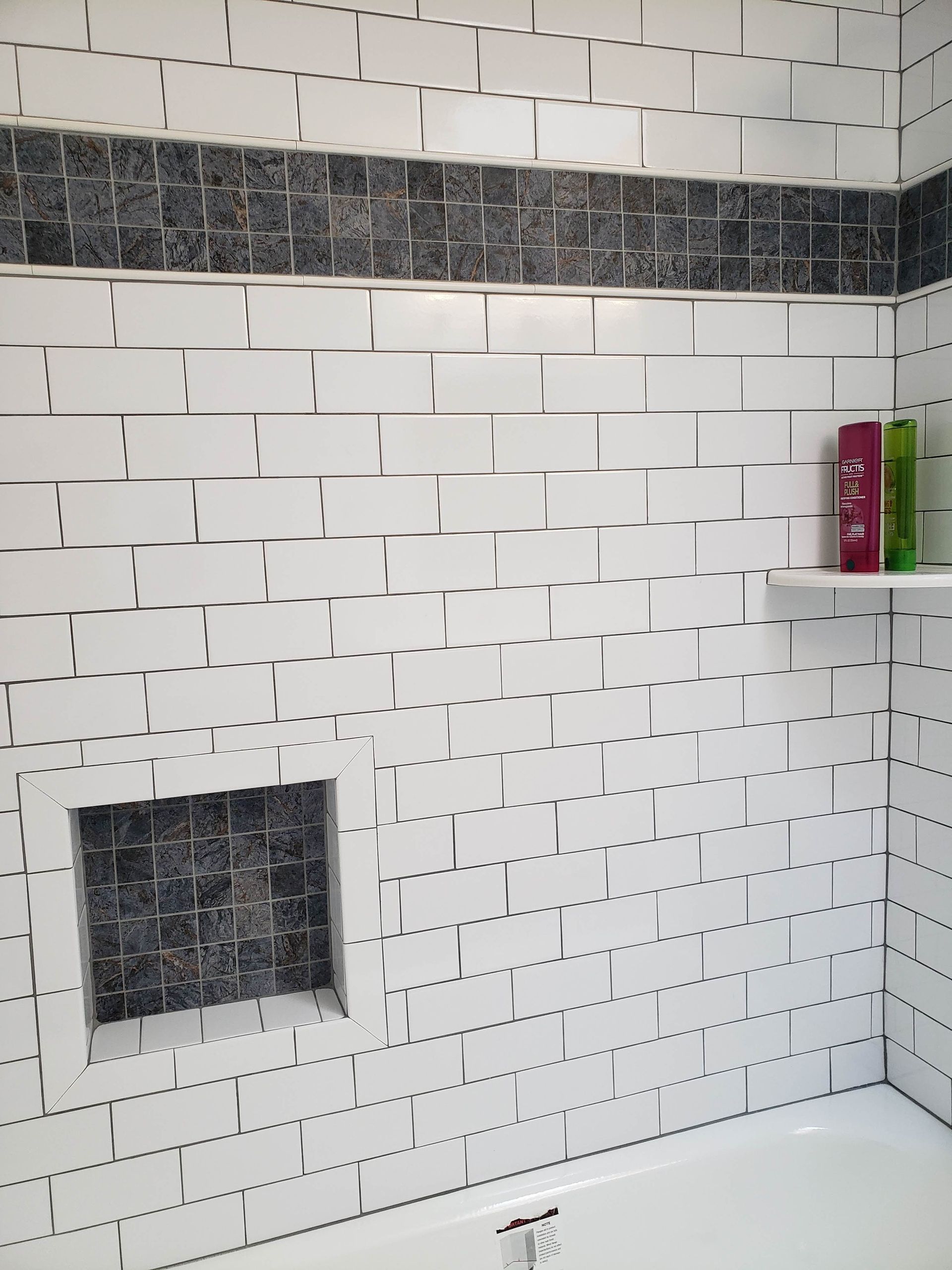 White tiled shower wall with dark mosaic accent band, recessed shelf, and bathtub corner