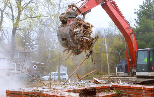 Red excavator demolishing a brick structure, holding debris in its claw; white vehicle and trees in the background.