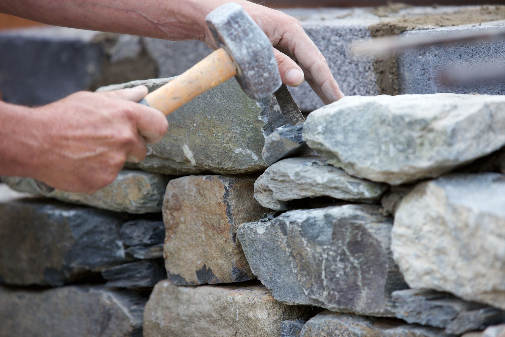 Hands use a stone mason's hammer to shape and place a piece of rock on a stone wall.