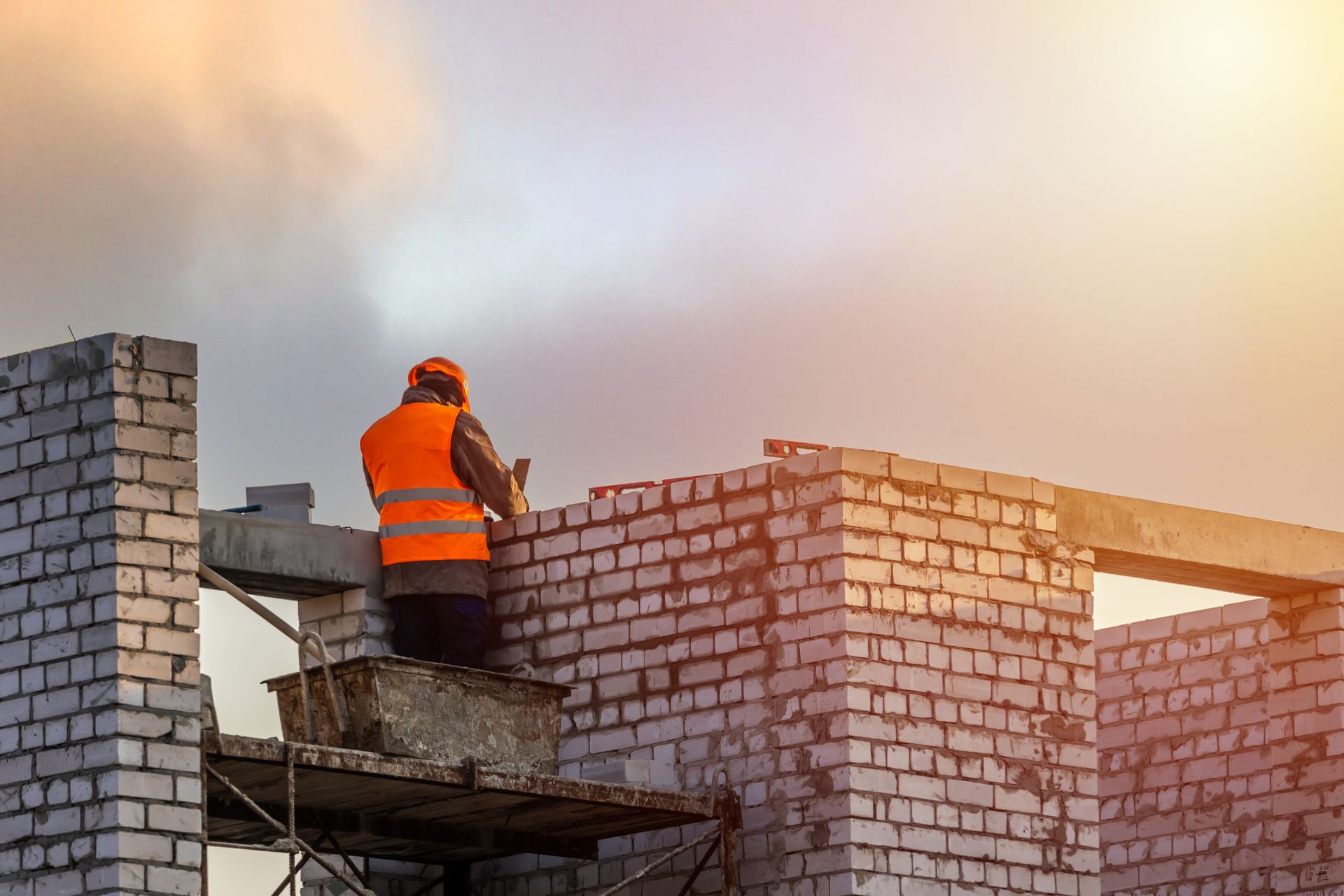 A worker in an orange safety vest building a white brick wall at a construction site against a bright.