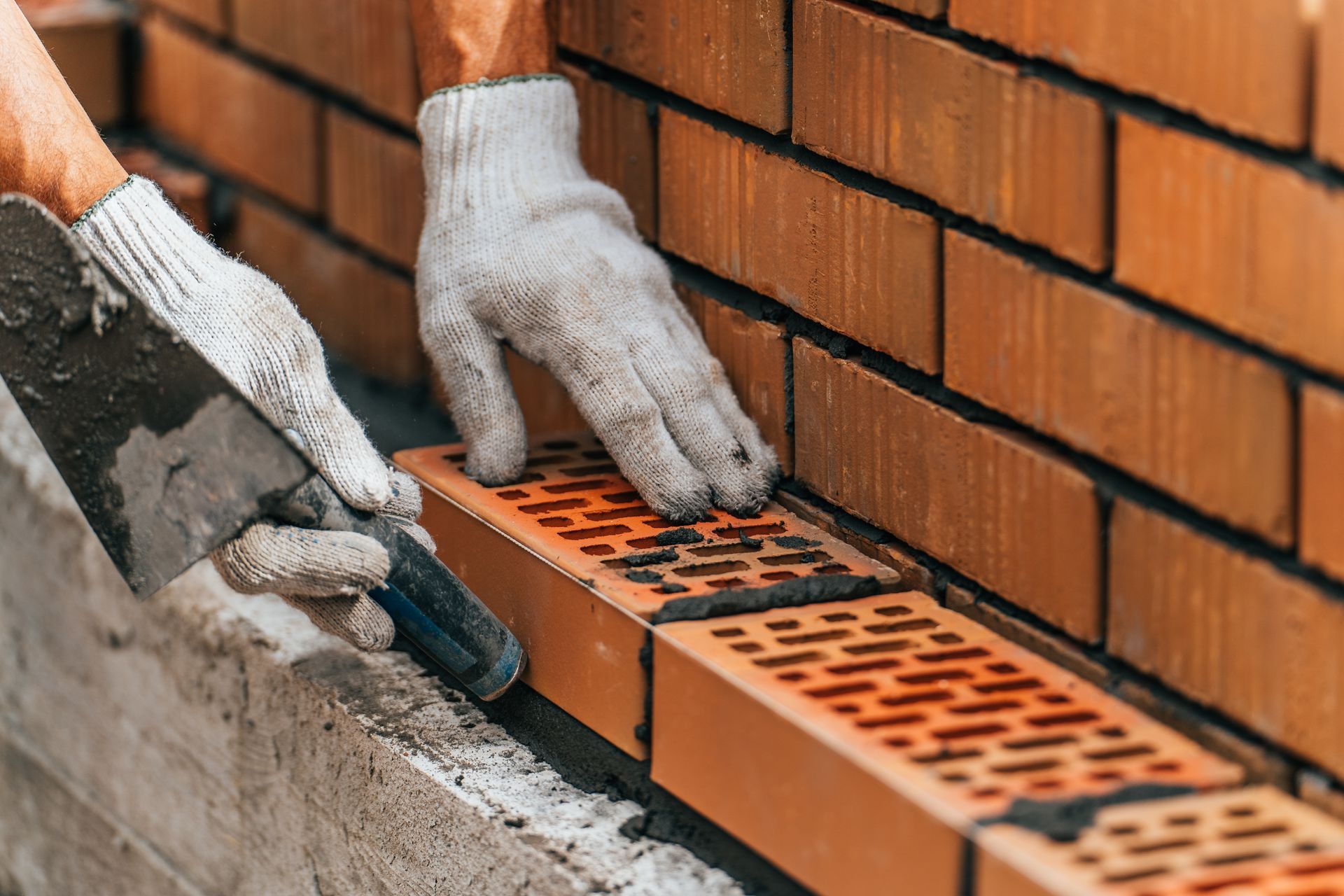 A worker wearing white gloves uses a metal trowel to spread mortar onto hollow red bricks while building a wall.