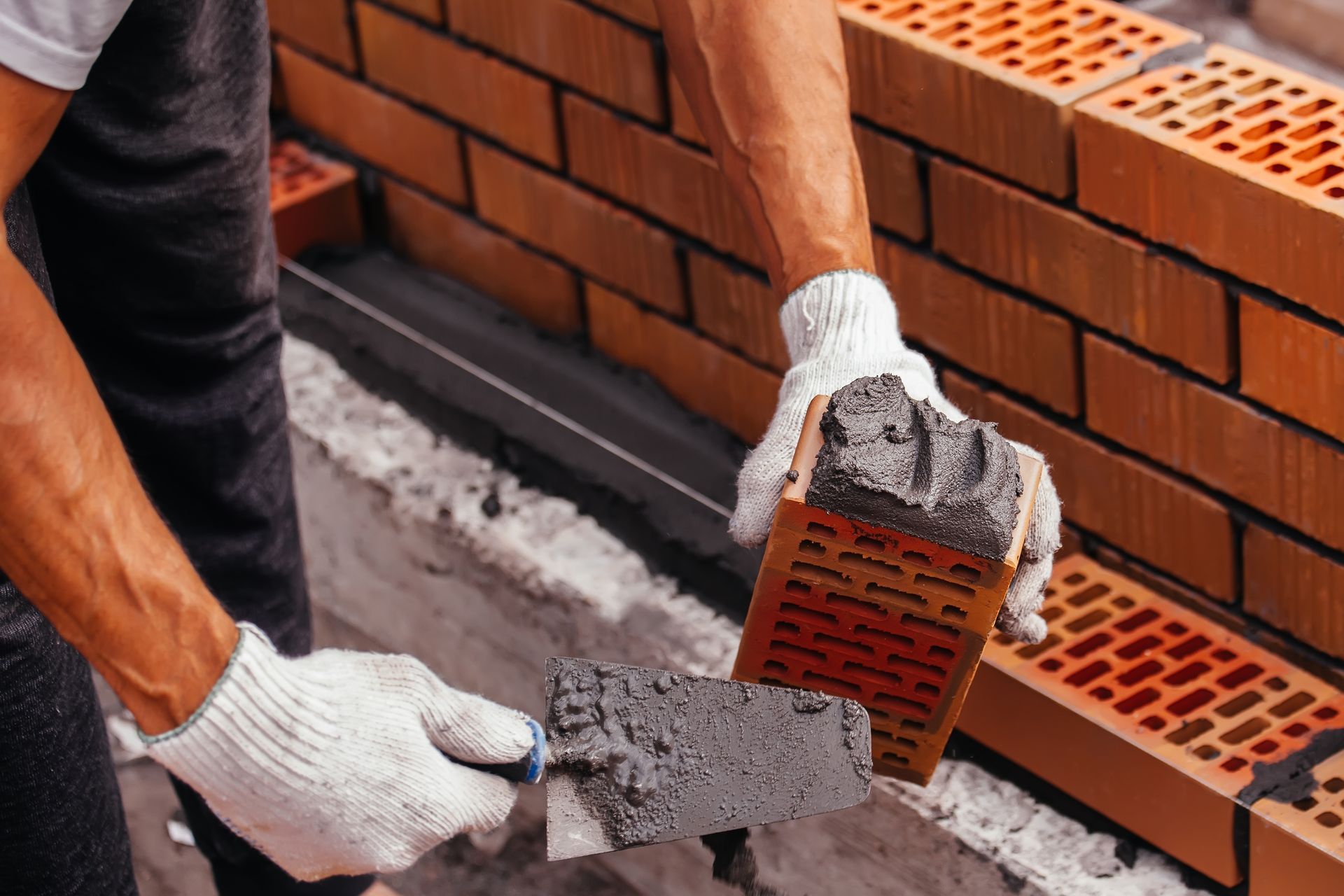 A mason wearing white gloves uses a trowel to apply mortar to a brick for building a wall.