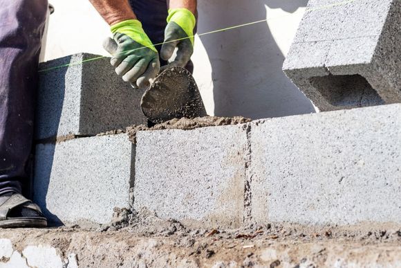 A worker in lime green gloves uses a trowel to apply mortar to gray concrete blocks being laid in a wall.