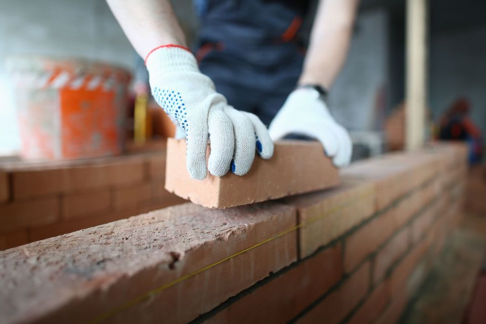A person wearing white work gloves carefully places a red brick onto a wall under construction.