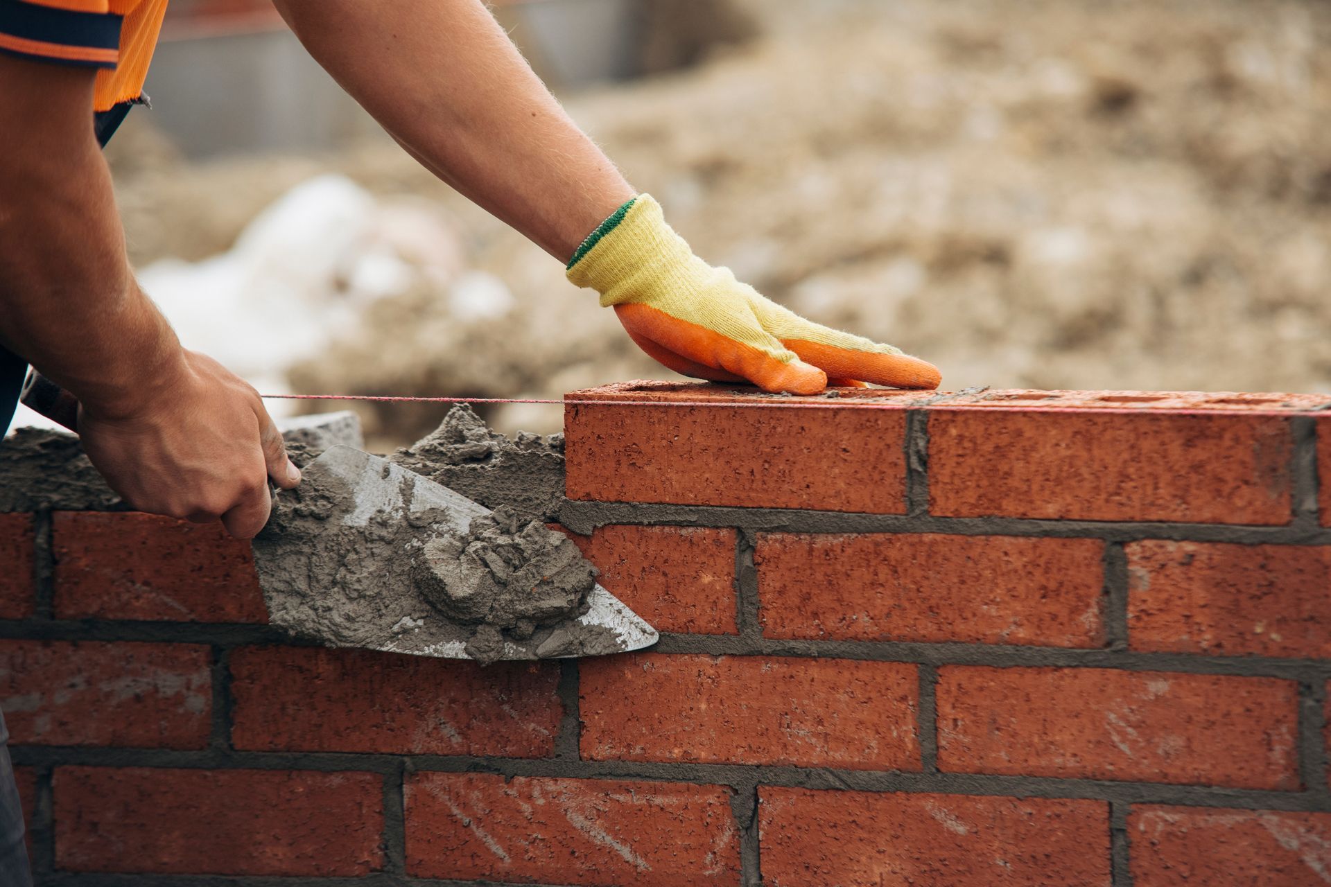 A worker wearing a yellow glove uses a trowel to apply mortar to a brick wall at a construction site.