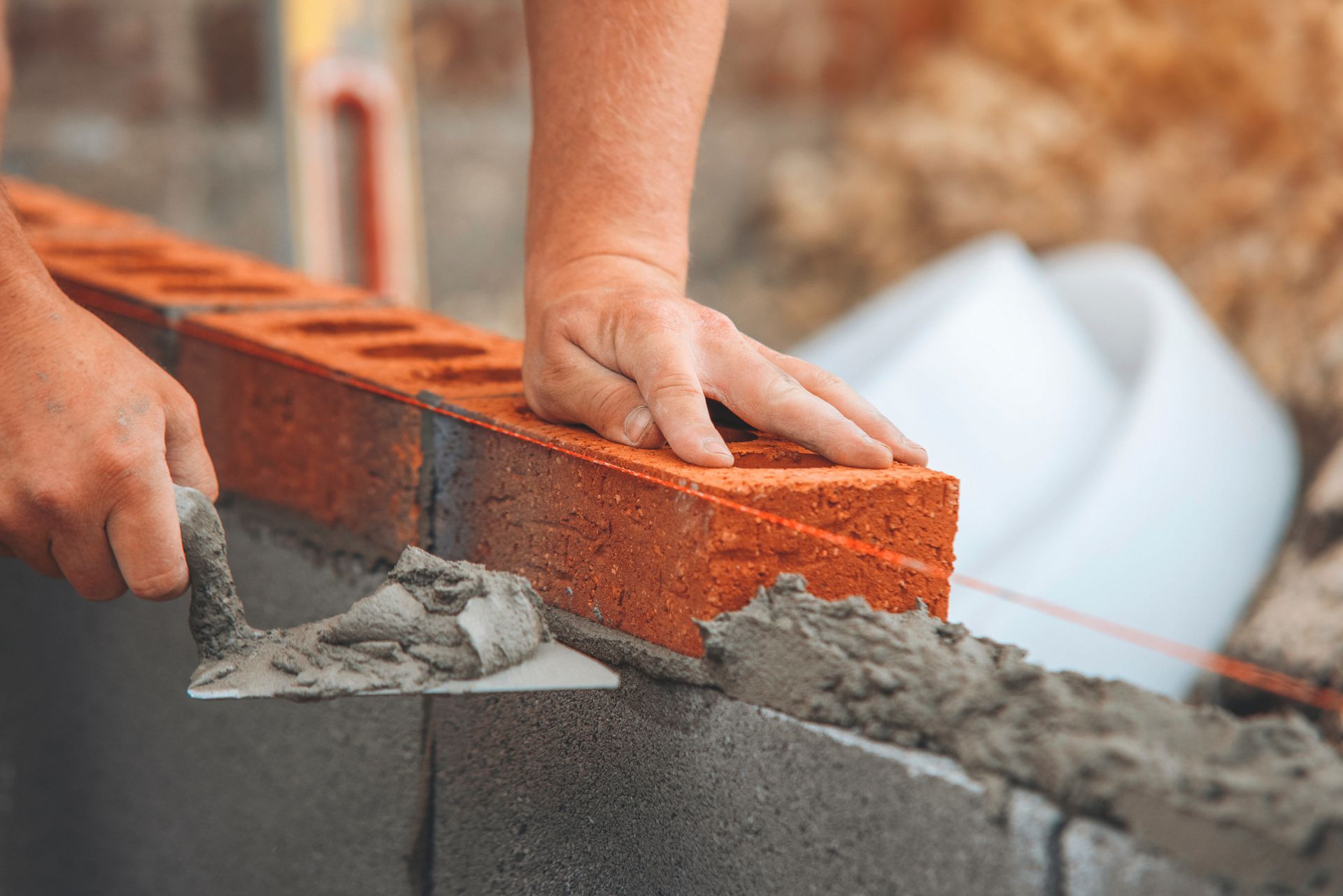 A close-up view of hands using a trowel to apply mortar between red bricks on a construction site.