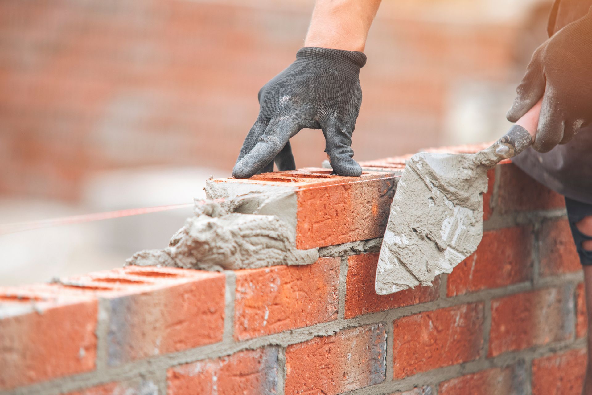 A close-up of a gloved hand using a trowel to apply mortar between red bricks during a construction project.