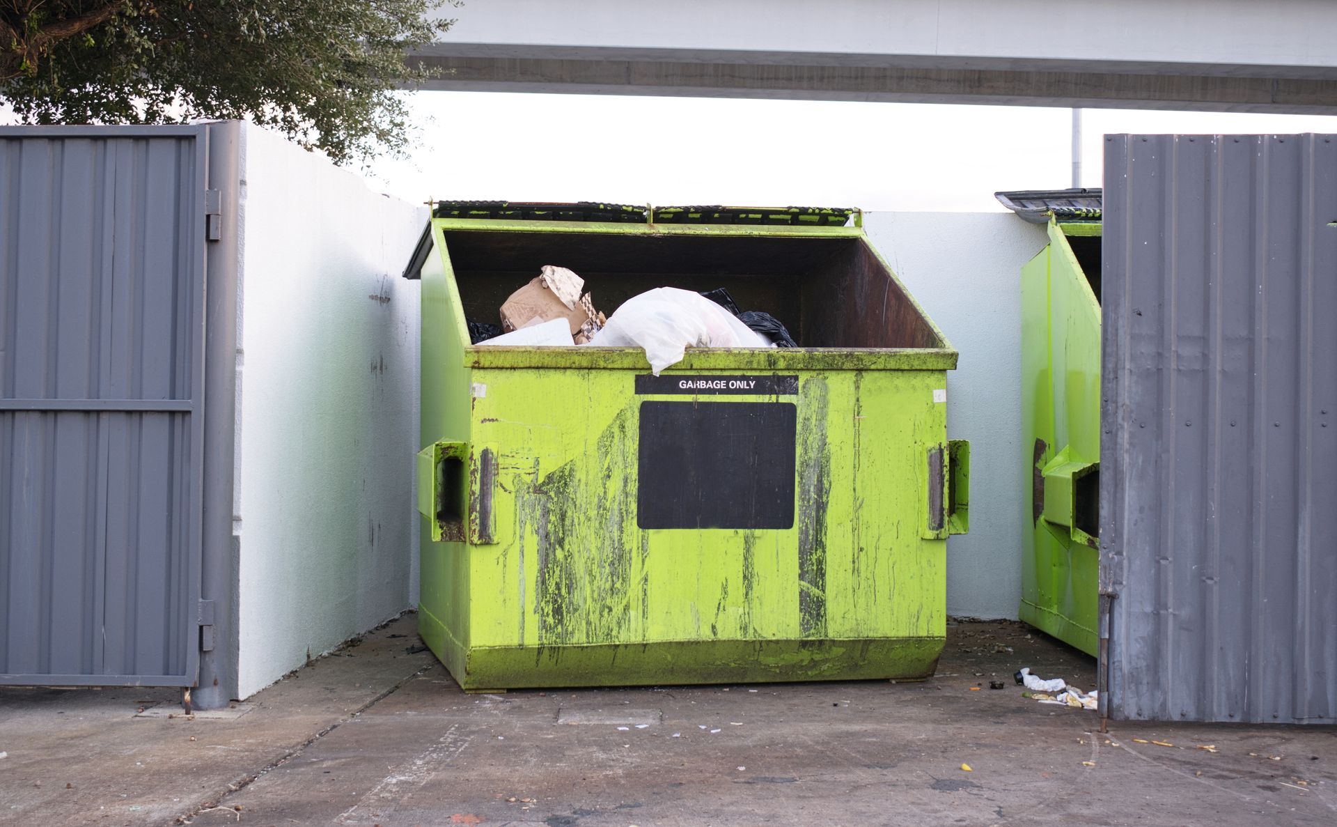 A weathered, neon-green dumpster sitting in an outdoor enclosure beneath a concrete overpass.