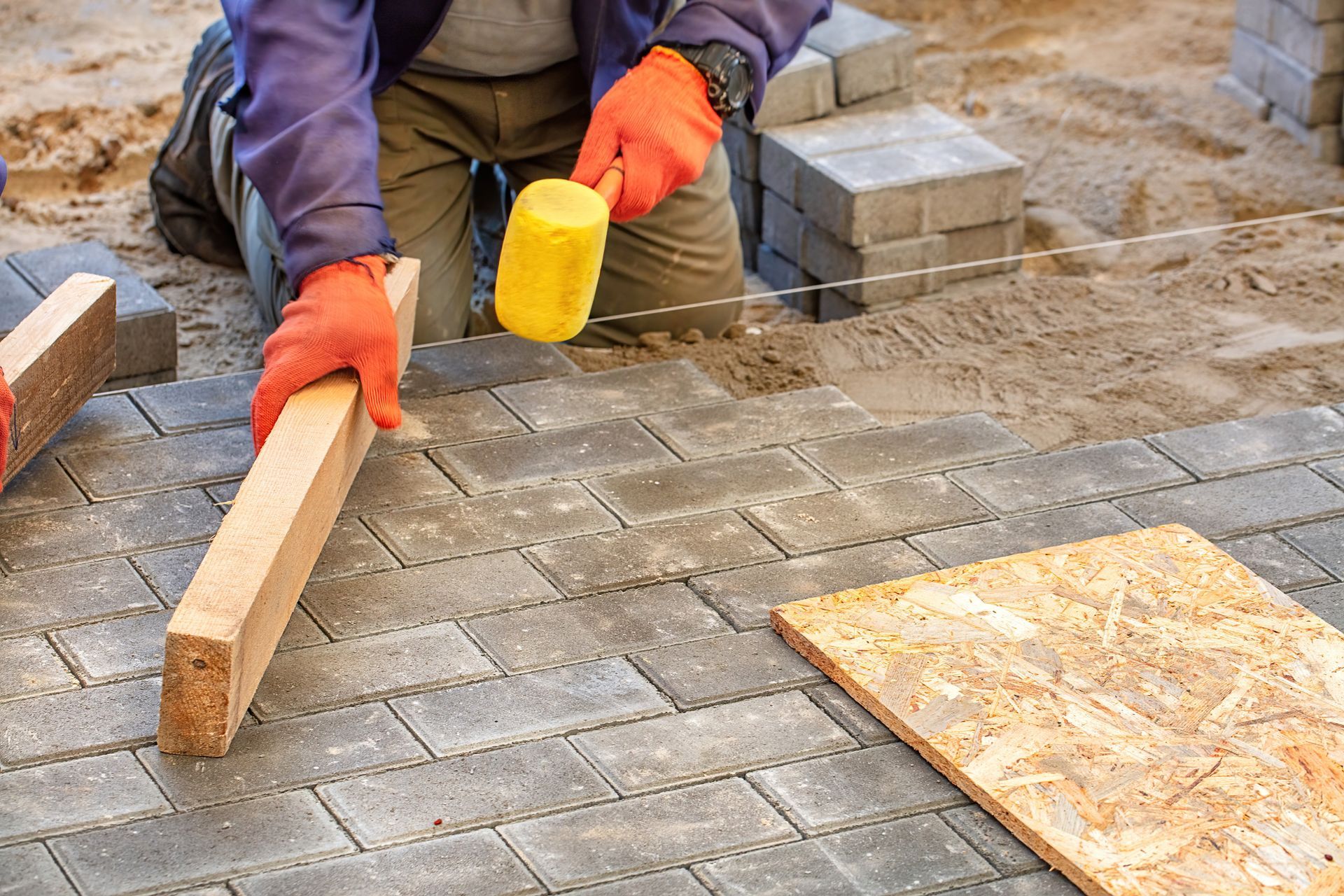 A person in orange gloves uses a wooden board and yellow mallet to level grey pavers on a sand construction site.