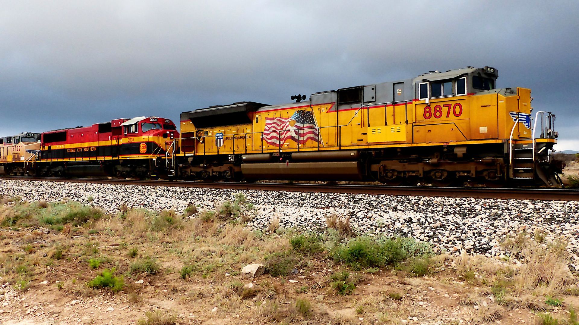 Locomotoras de carga amarillas y rojas arrastrando vagones por las vías en un paisaje seco y abierto.