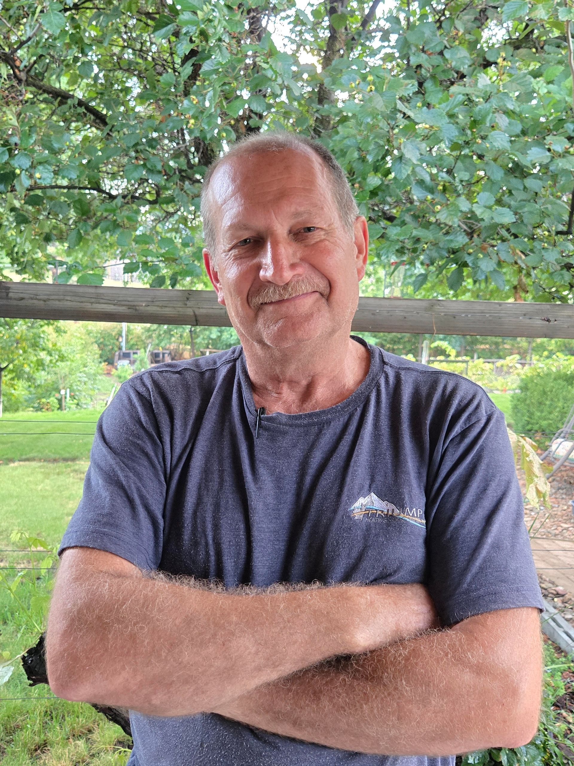 Smiling man with arms crossed in a blue T-shirt, sitting outdoors under leafy trees.