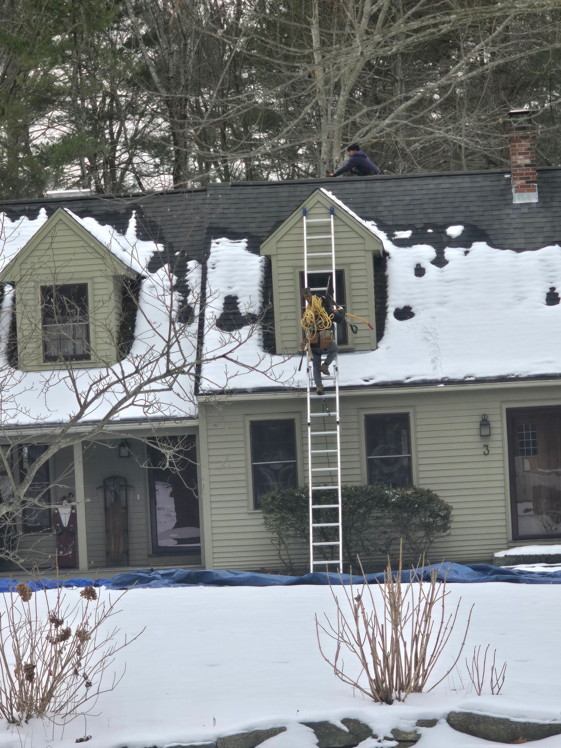Two-story house with snow-covered roof; person on ladder cleaning an upstairs window.