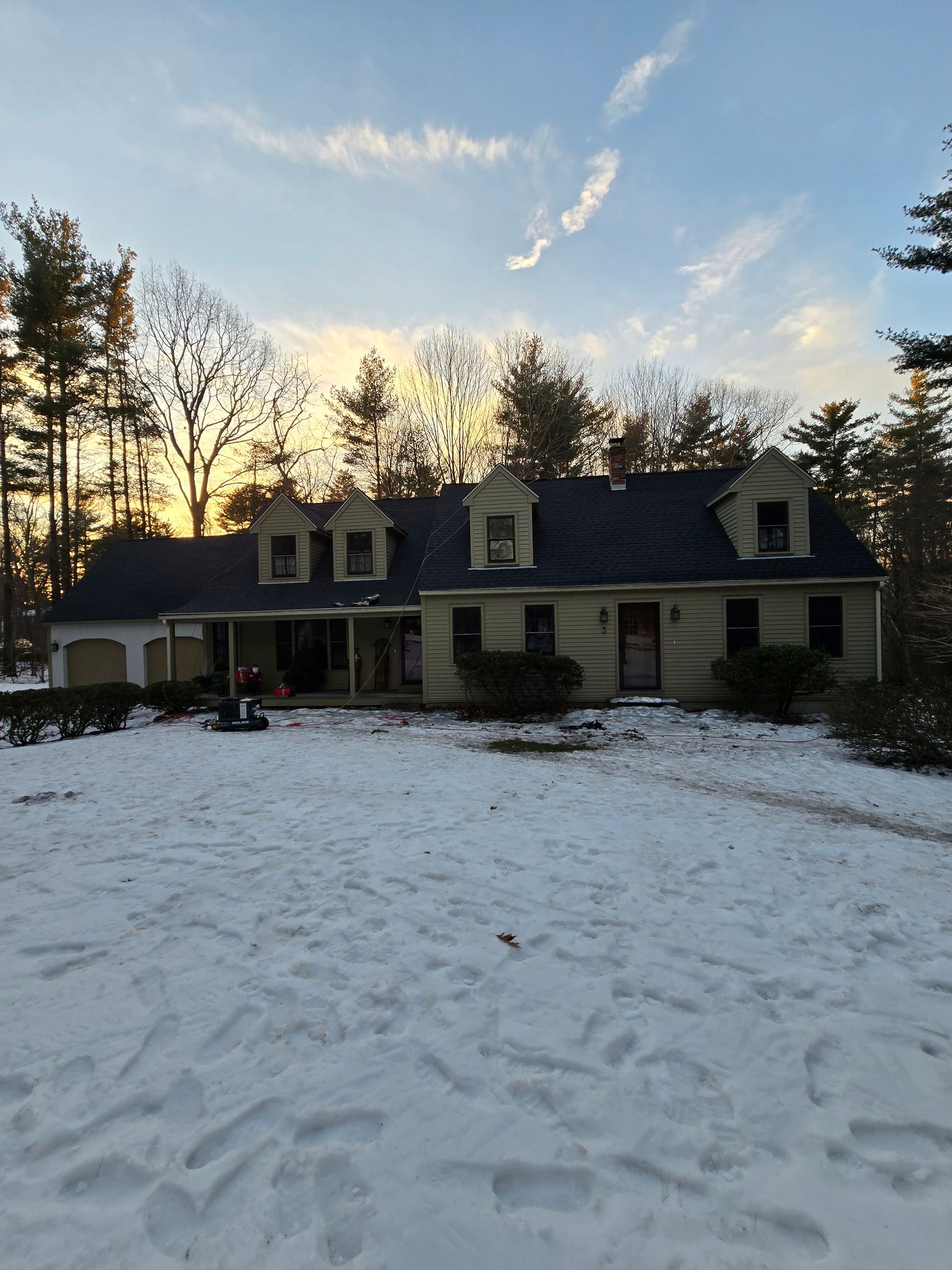 Snow-covered suburban house at sunset with footprints in the yard and bare trees silhouetted against a blue sky