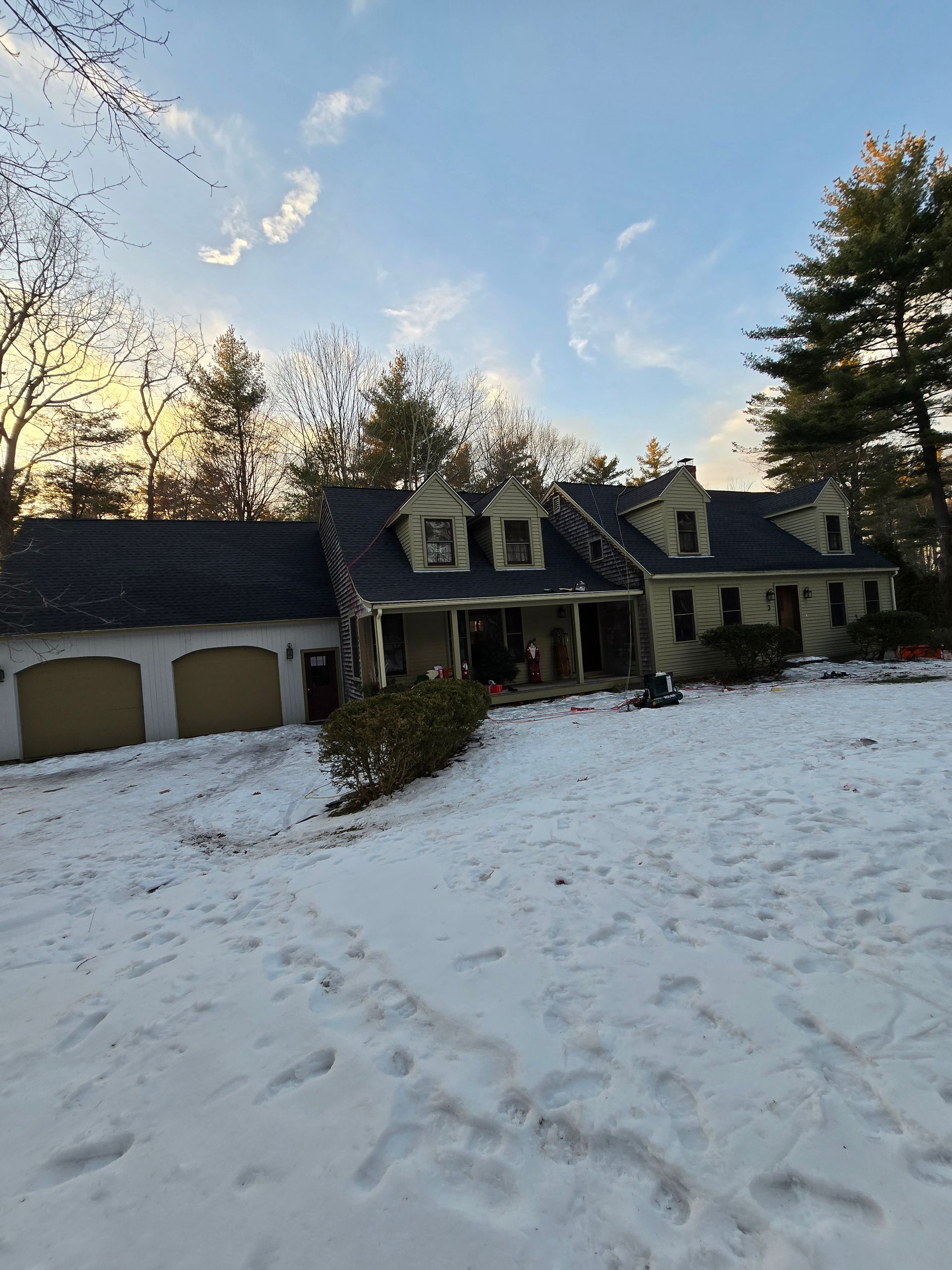 Snow-covered suburban house with a porch, garage, and footprints in the front yard at sunset.