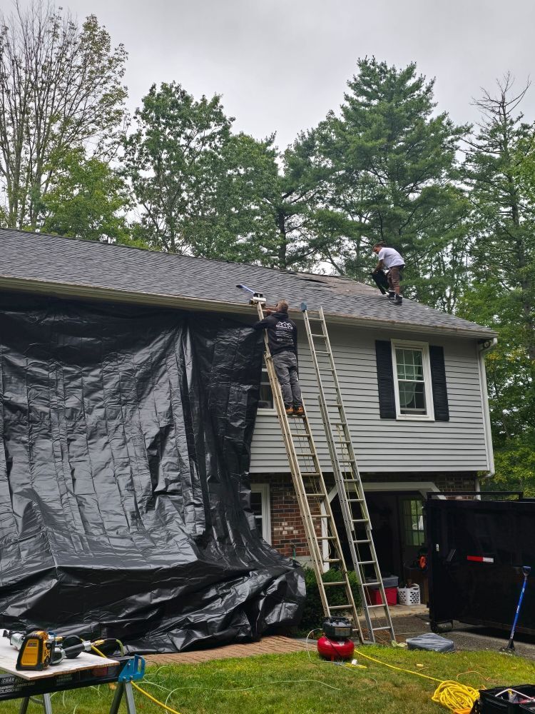 Two workers on a roof, one on a ladder, black tarp covers side of house, cloudy day.
