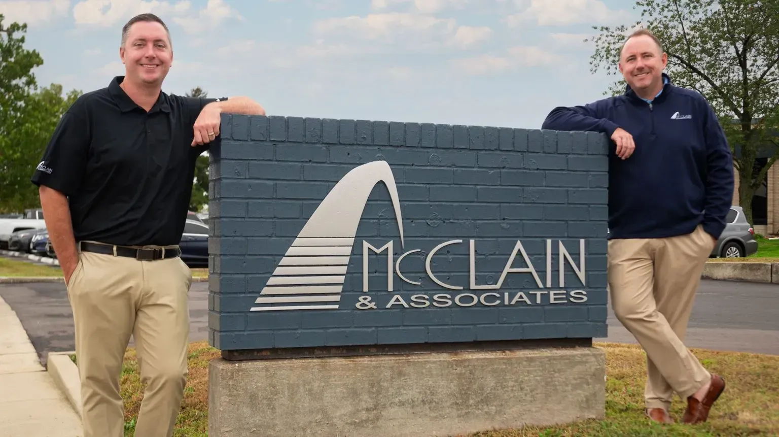 Two men posing beside a McClain & Associates sign outdoors on a cloudy day