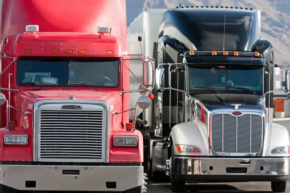 Two semi trucks, one red and one silver, parked side by side, facing forward.