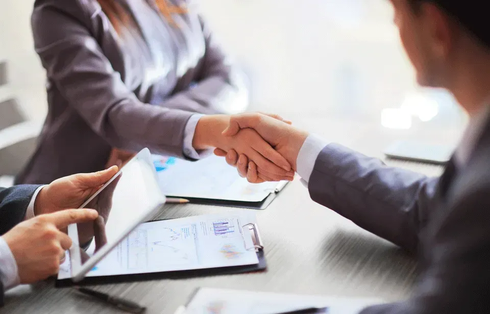 Two businesspeople shaking hands across a table with documents and a tablet nearby