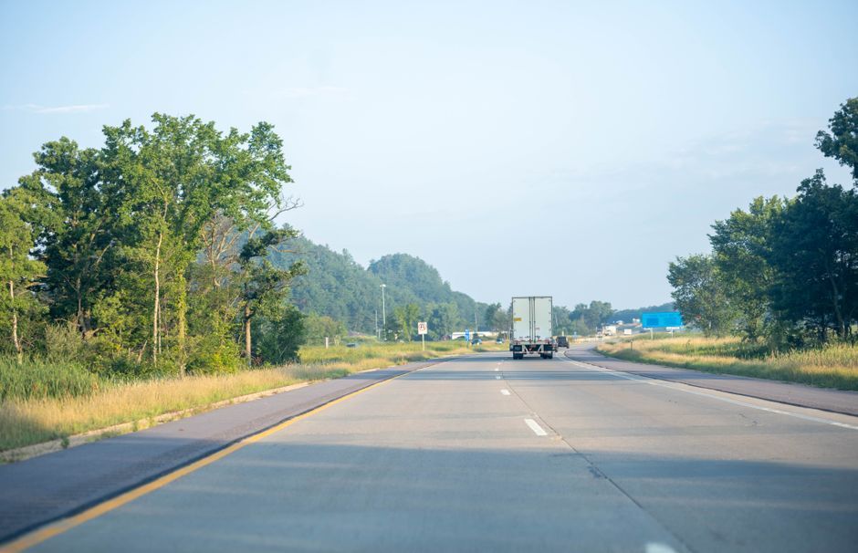 Highway view with a truck ahead, trees lining the road, and a clear blue sky.