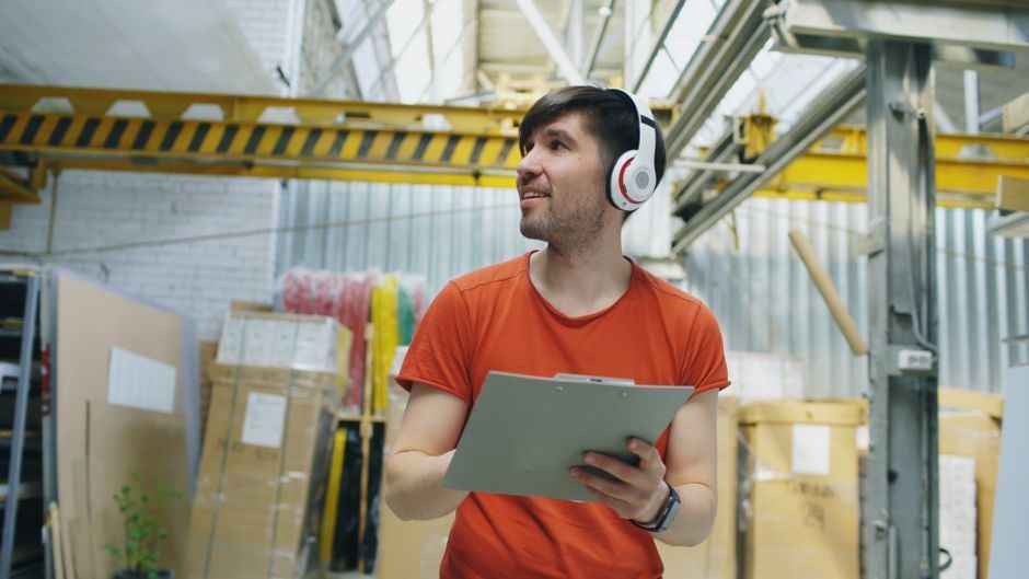 Warehouse worker in an orange shirt and headset holding a clipboard and looking up among stacked boxes