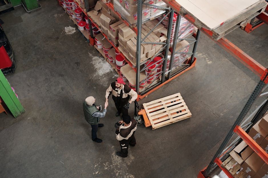 Three workers stand beside stacked pallets in a warehouse aisle, viewed from above.