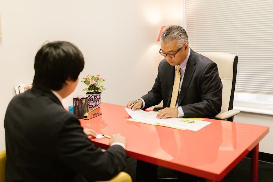 Two people in suits meeting at a red table, one writing on paper in a small office.